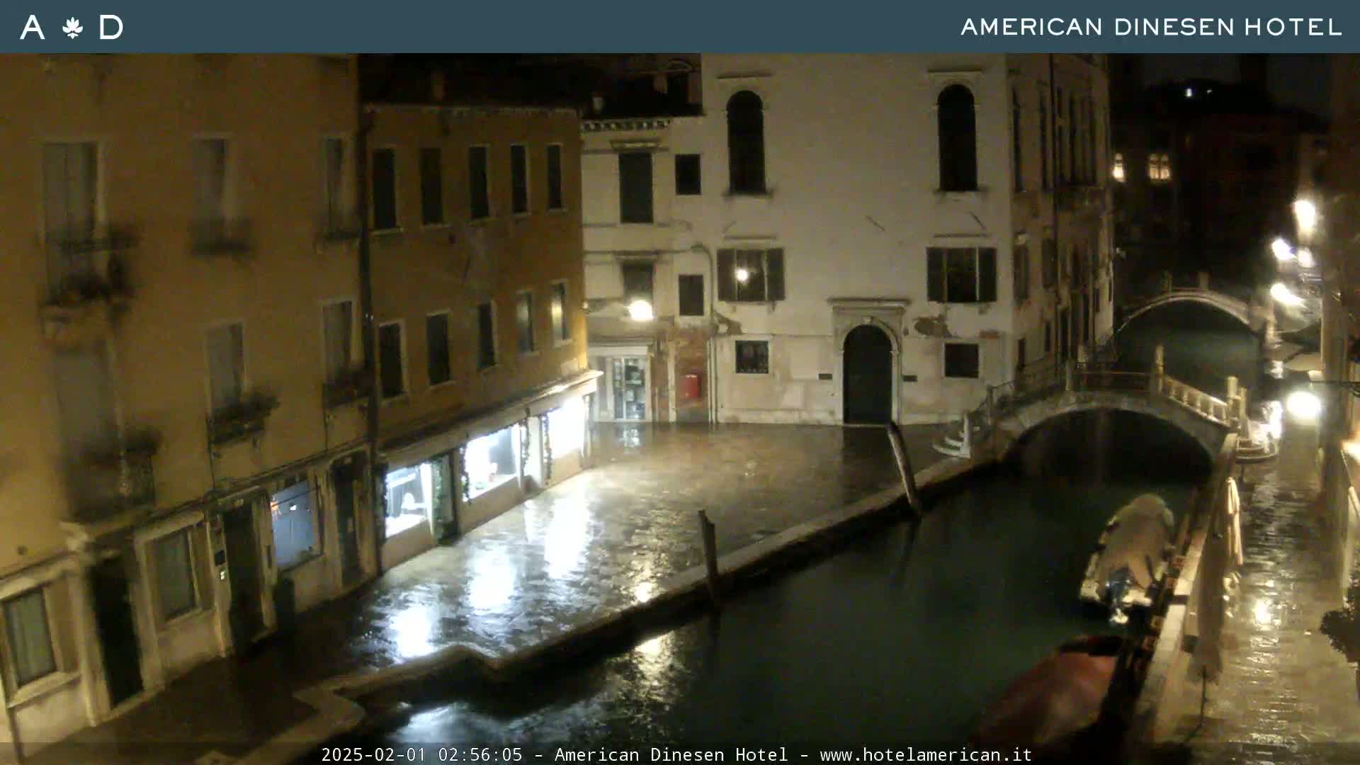 A nighttime view of a canal in Venice, showing several buildings and a bridge, with wet ground indicating rain.