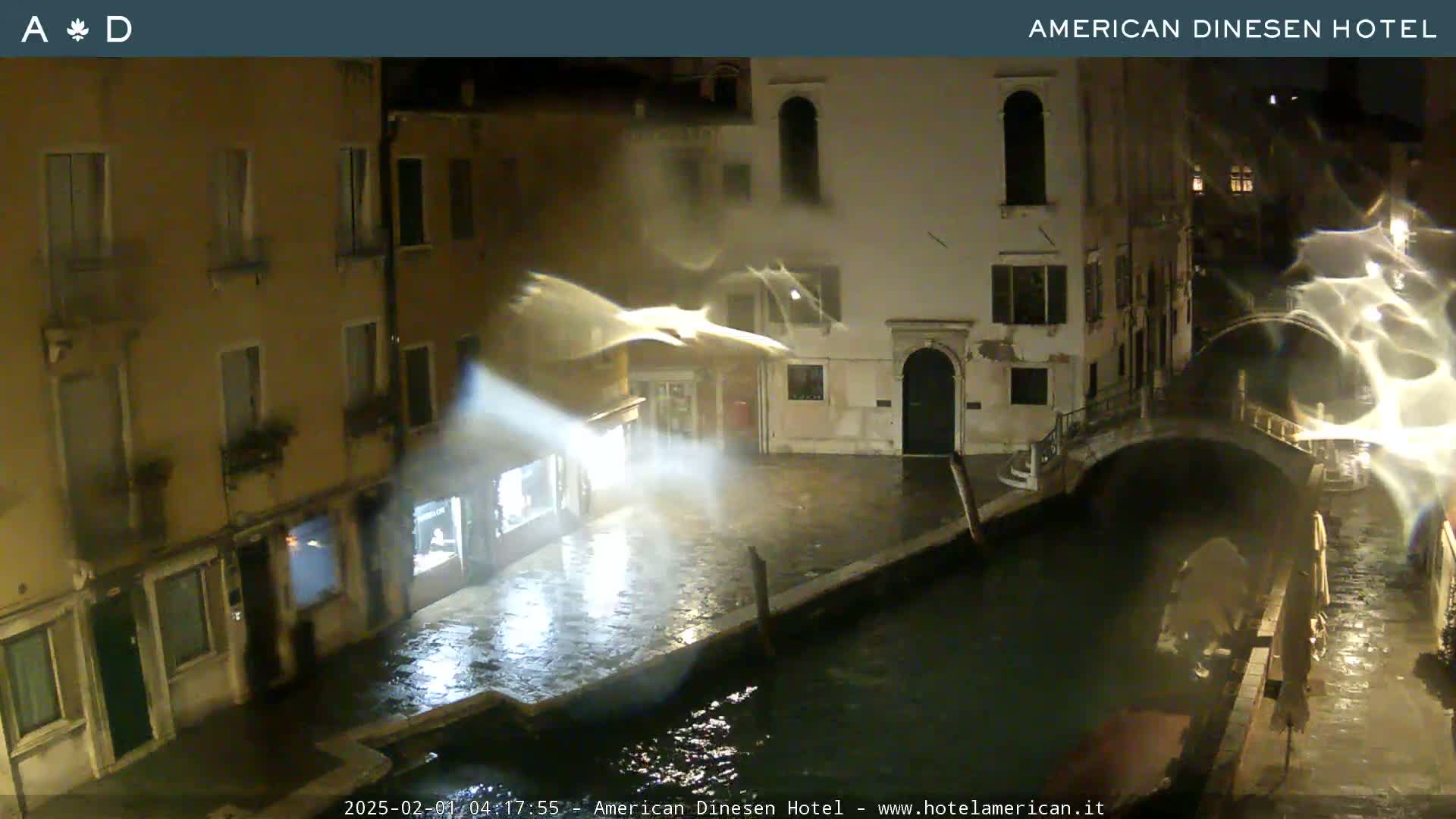A nighttime view of a canal in Venice, Italy, showing rain-slicked streets and buildings, with a bridge arching over the water.