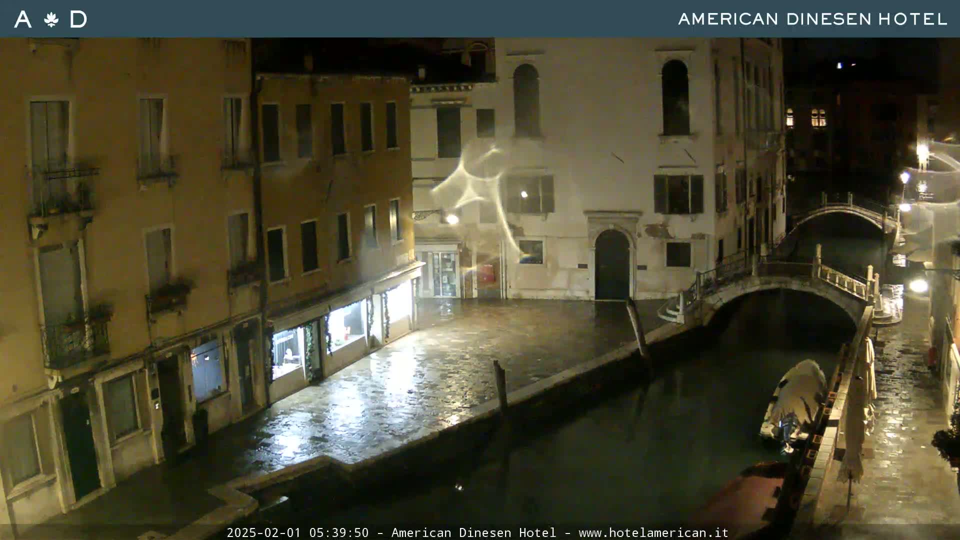 A nighttime view of a canal in Venice, Italy, showing wet cobblestone streets and buildings, with a small boat moored alongside a bridge under a light rain.