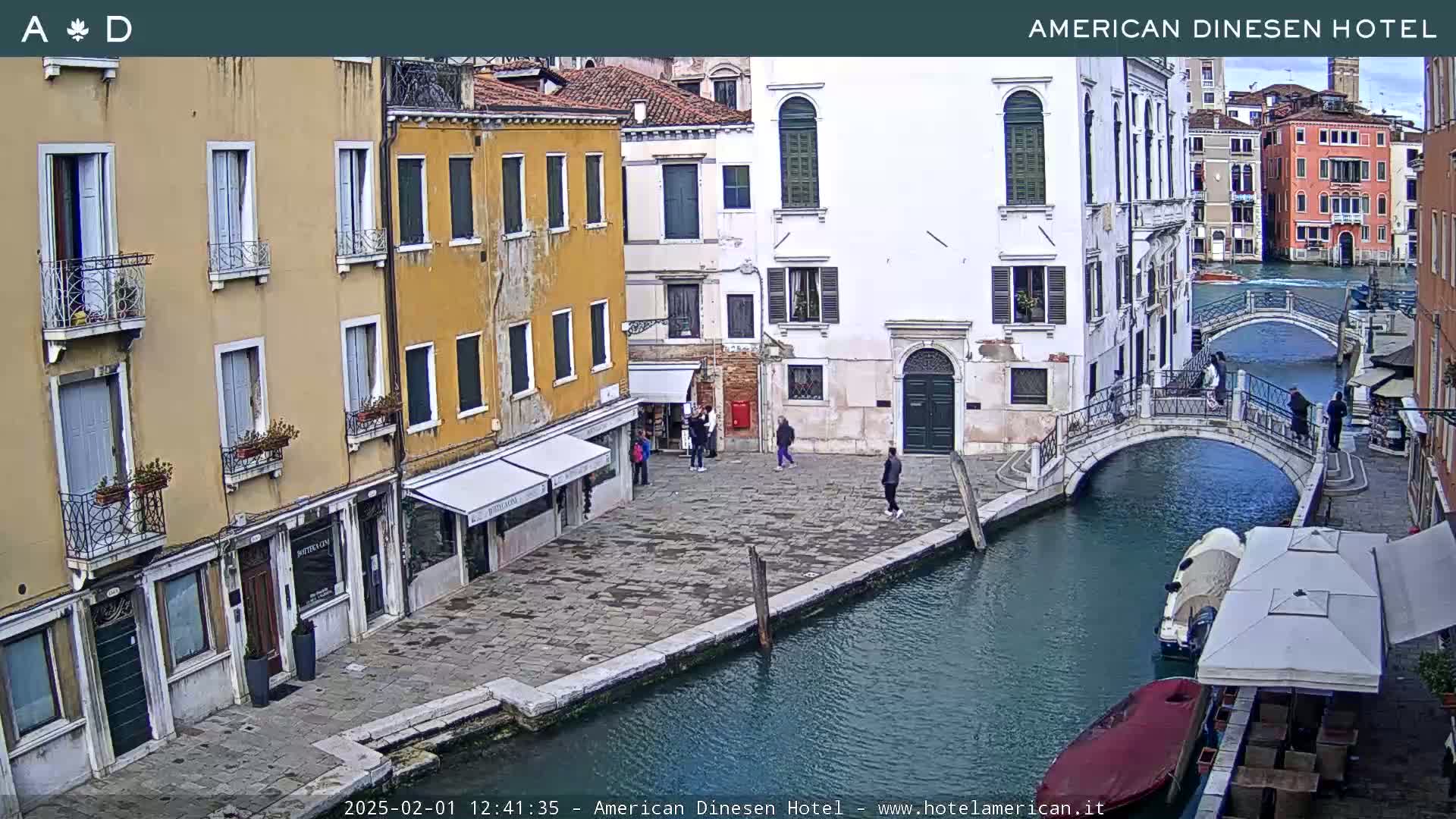 A canal in Venice, Italy, with several buildings lining its banks, a small bridge spanning the waterway, and a few people walking along the edge on a seemingly clear day.