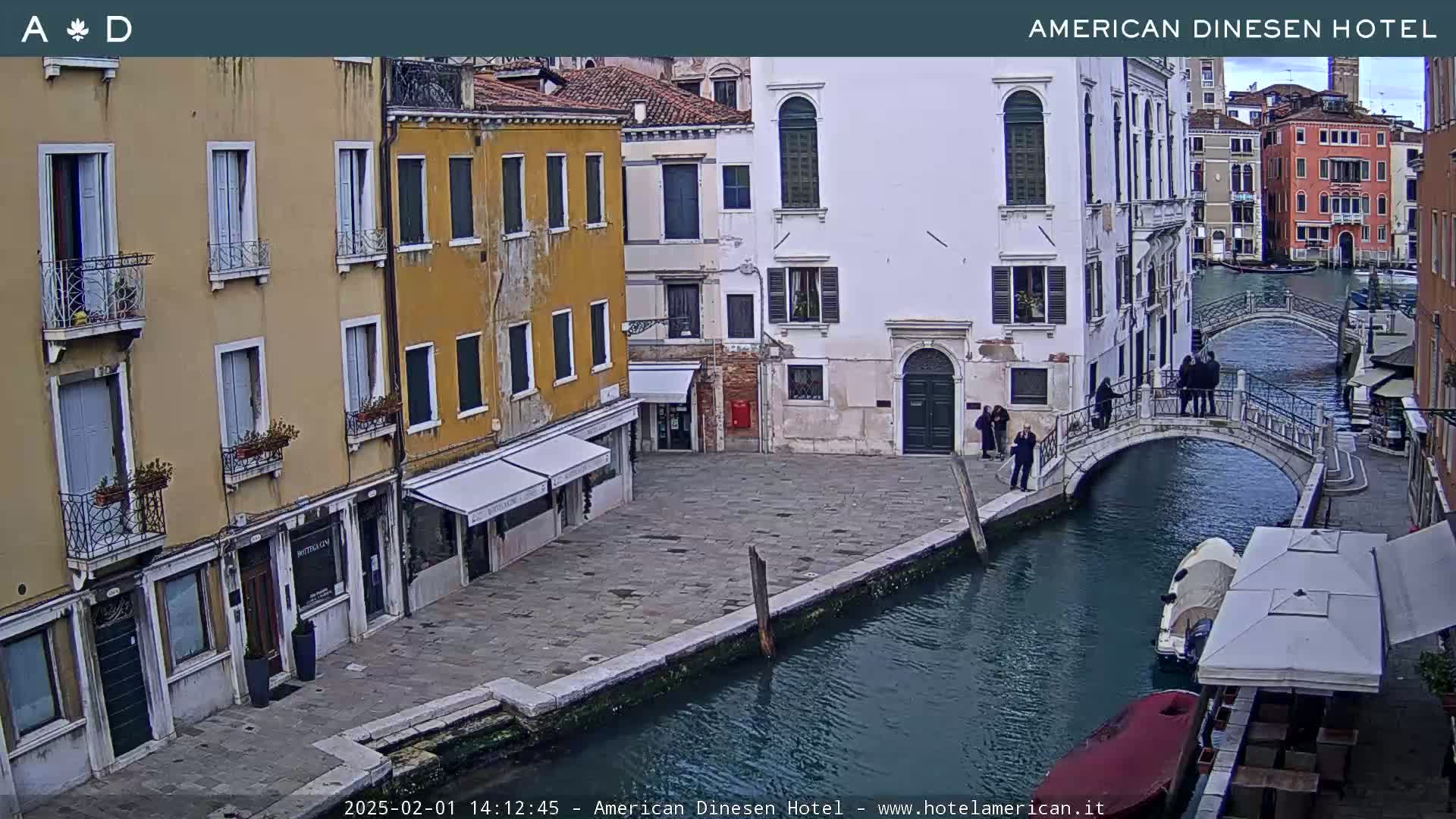 A canal in Venice, Italy, with several multi-story buildings lining its banks, a small footbridge spanning the waterway, and a few people walking across it on an overcast day.