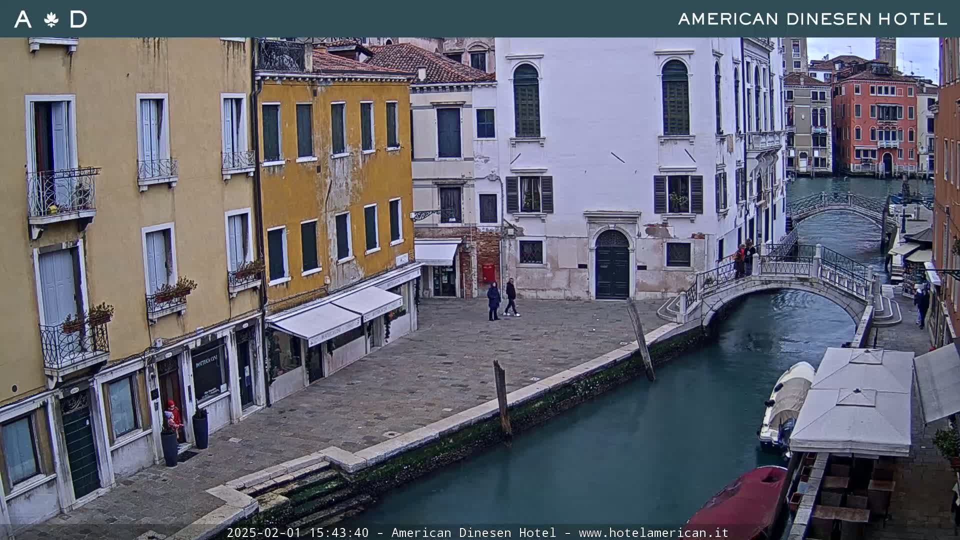 A canal in Venice, Italy, with several buildings lining its banks, a small bridge crossing it, and a few people walking along the waterway, under an overcast sky.
