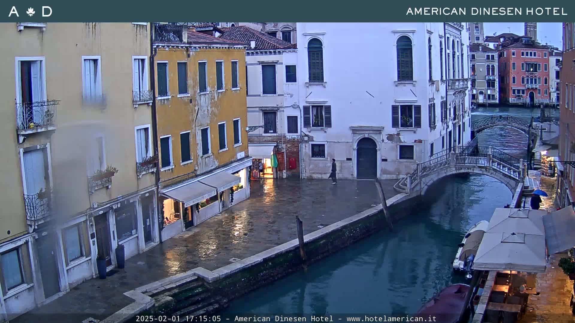 A rain-slicked canal in Venice, lined with tall, narrow buildings, is spanned by a small bridge, with a boat moored alongside a cafe.