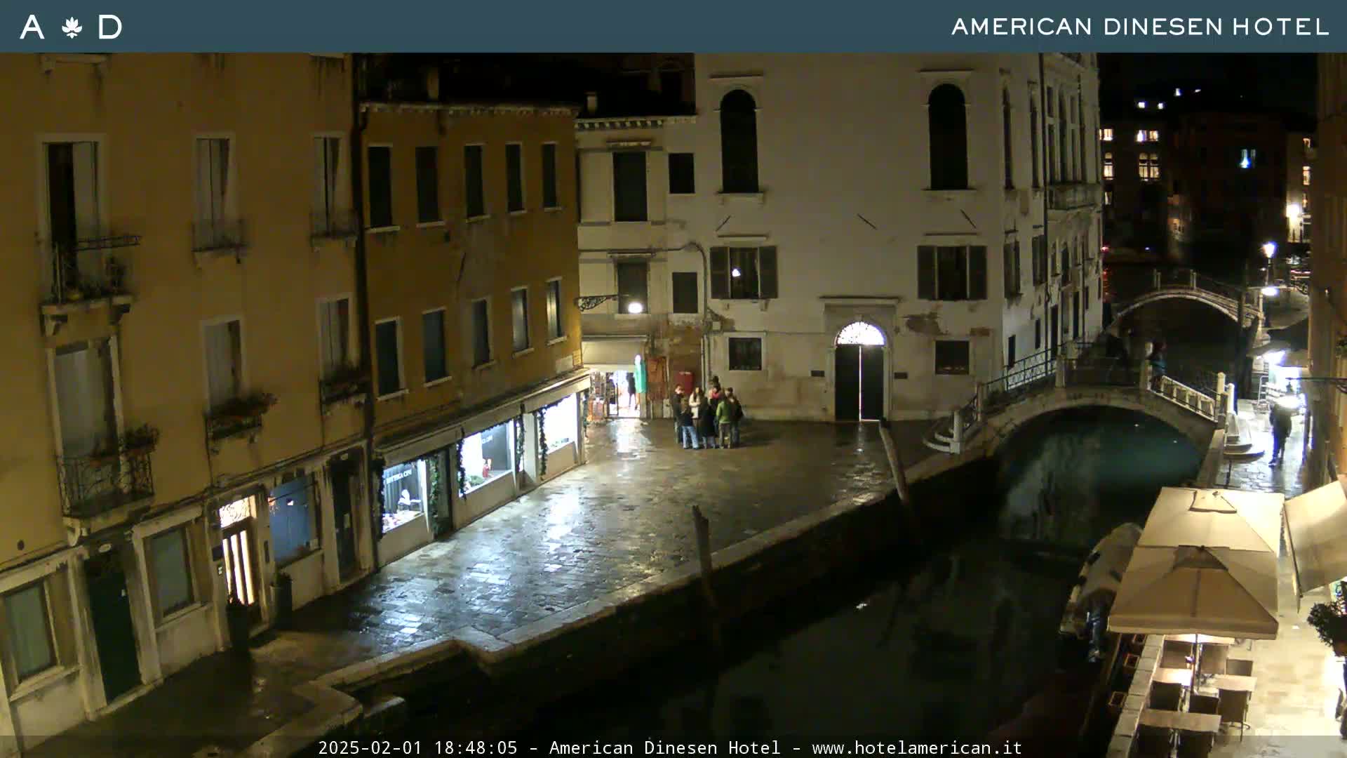 A nighttime view of a canal in Venice, with several buildings lining the waterway, a small group of people standing near a building, and an outdoor patio area with umbrellas.