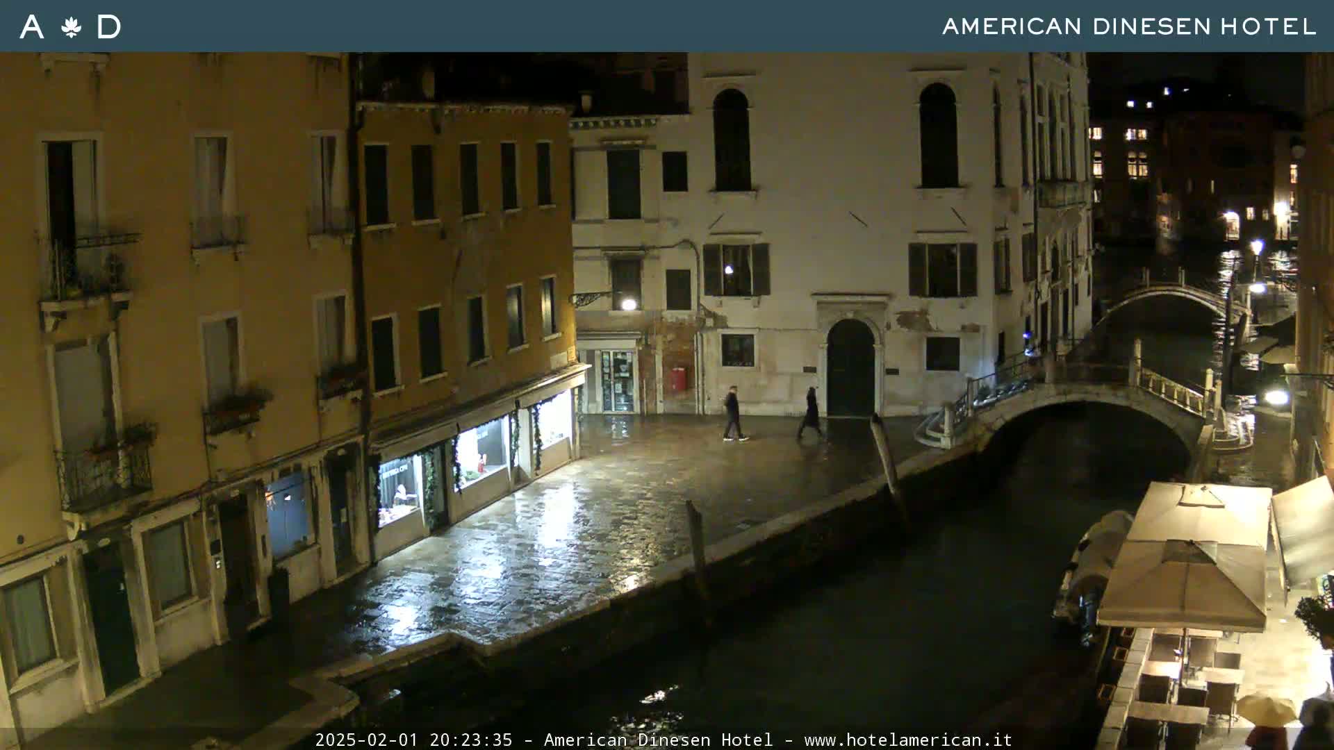 A nighttime view of a canal in Venice, Italy, shows wet cobblestone streets and buildings lining the waterway under a dark sky.