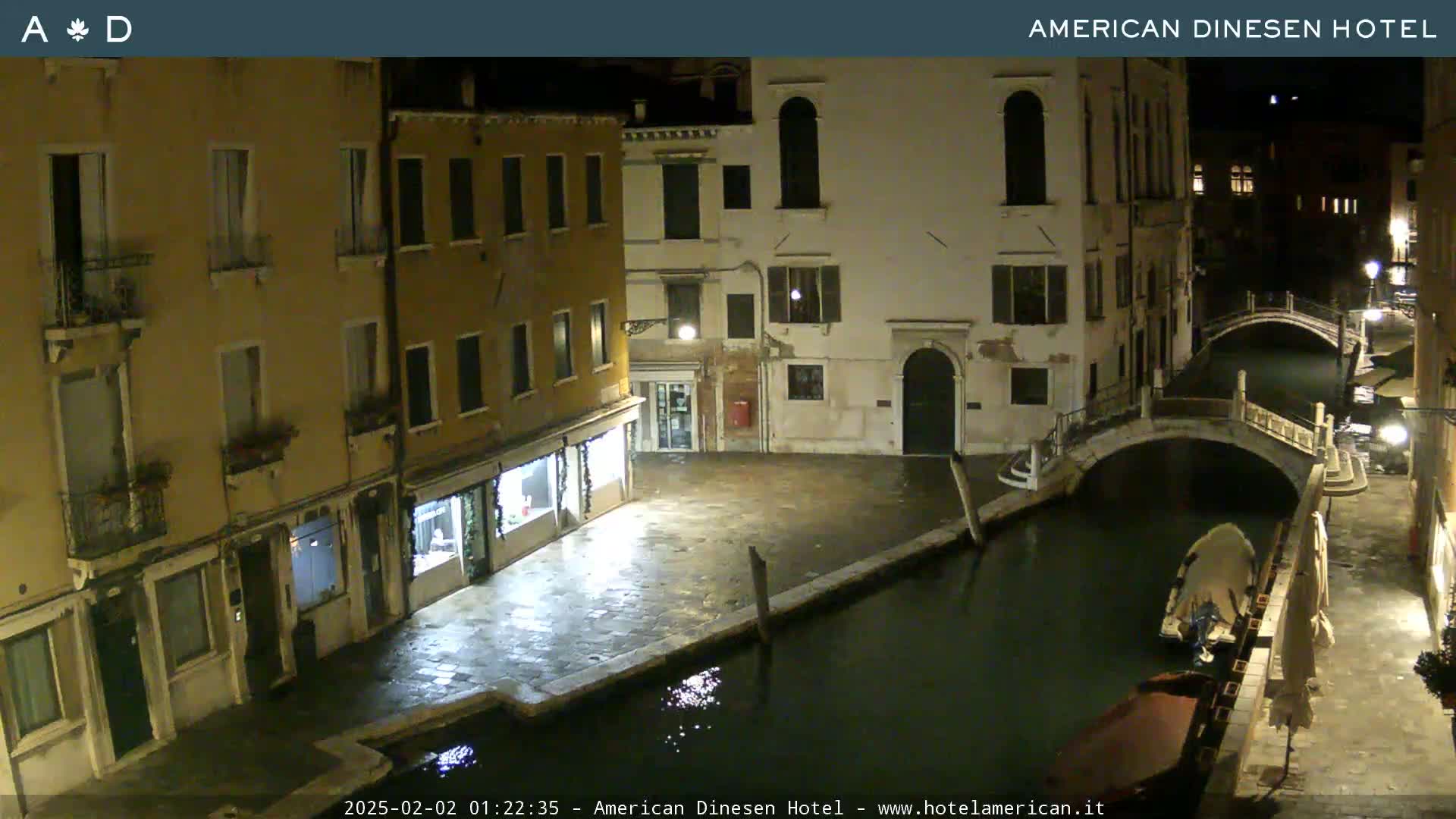 A nighttime view of a canal in Venice, Italy, shows several buildings lining the waterway, with a small boat moored alongside a low bridge.
