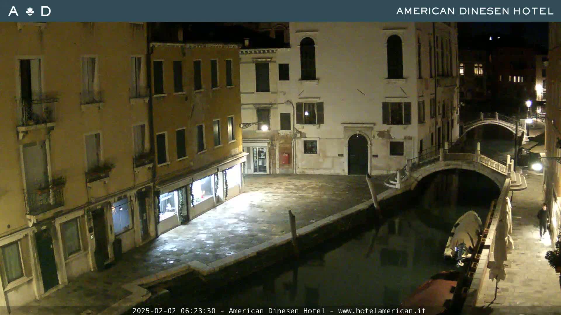 A nighttime view of a canal in Venice, showing several buildings along the waterway and a small bridge, with a boat moored alongside.