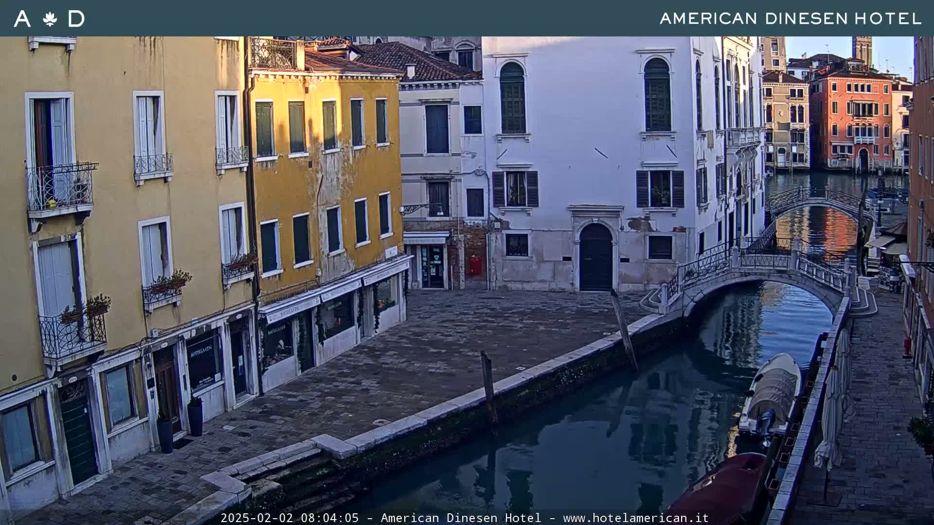 A tranquil canal scene in Venice shows several buildings lining the waterway, with a small bridge and a boat on the water under a clear, early morning sky.