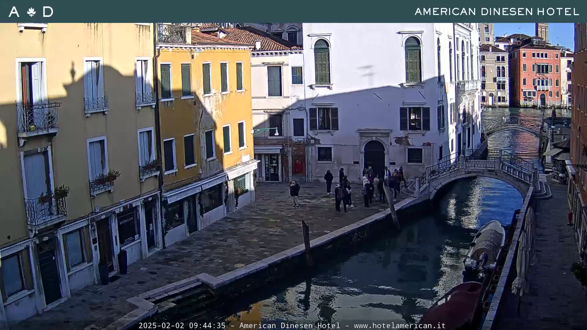 A sunny view of a canal in Venice, Italy, shows several buildings lining the waterway, a small bridge spanning the canal, and a group of people standing on the canal's edge.