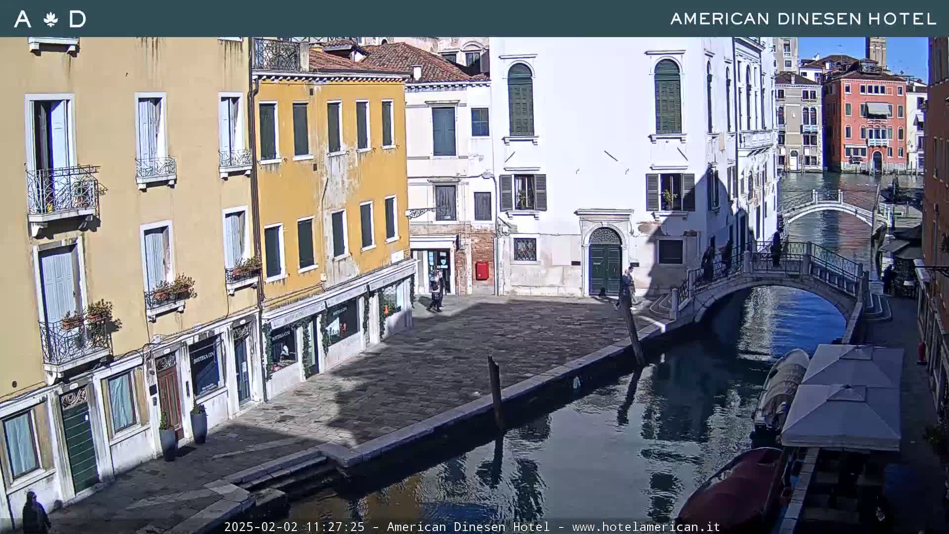 A sunny Venetian canal scene shows several buildings lining the waterway, a small bridge crossing the canal, and a few pedestrians.