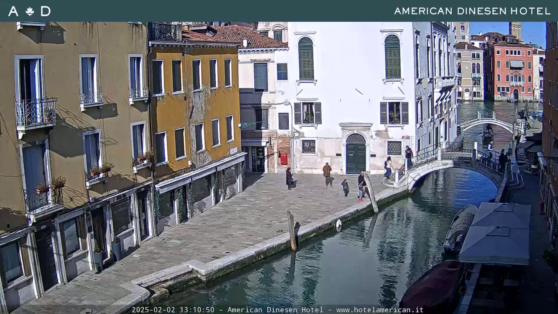 A sunny view of a canal in Venice, Italy, shows several buildings lining the waterway, a small bridge, and a few people walking along the canal's edge.