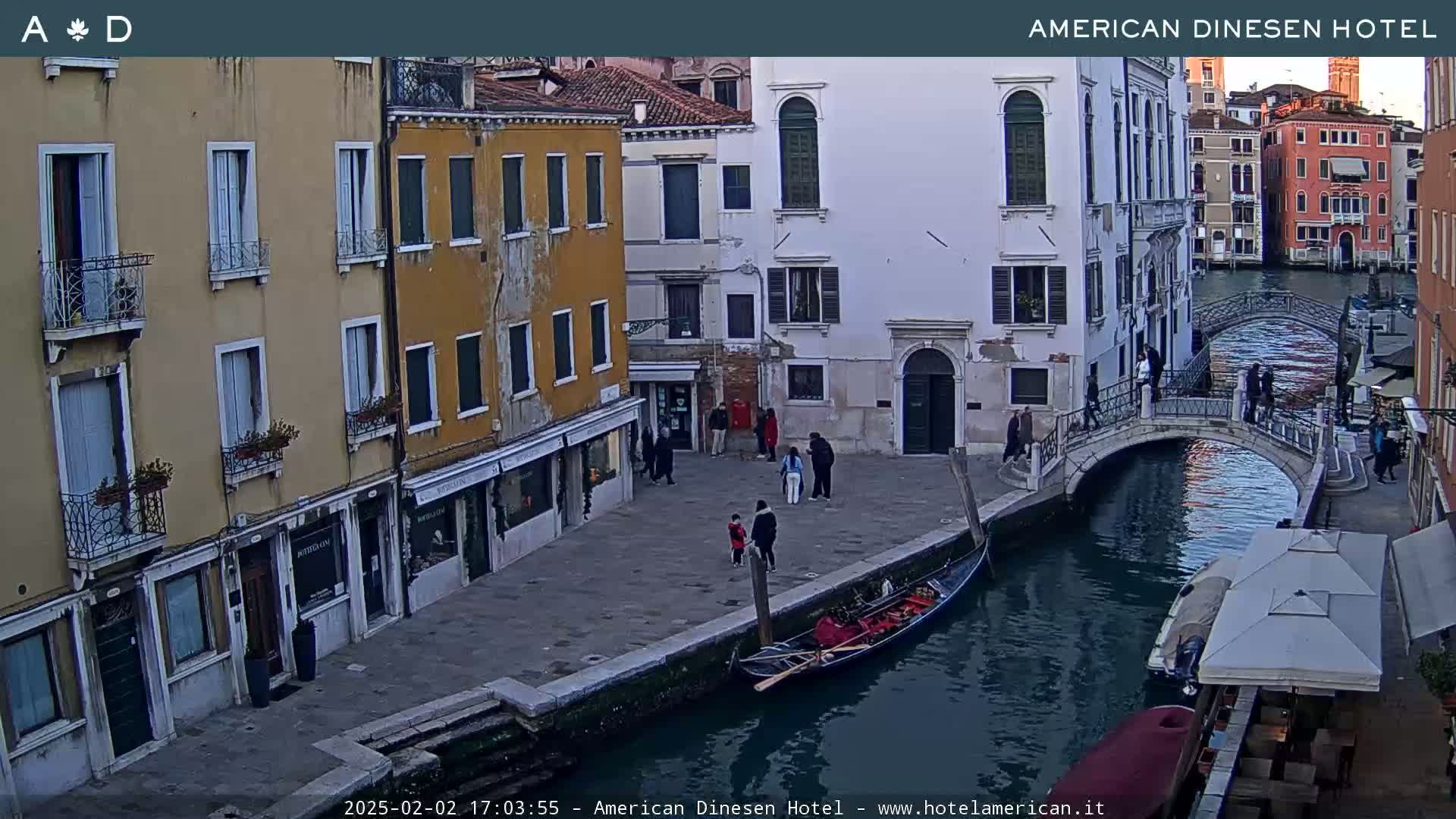 A gondola is moored along a canal in Venice, Italy, under a partly cloudy sky, with several people walking along the canal's edge and a small bridge crossing the waterway.