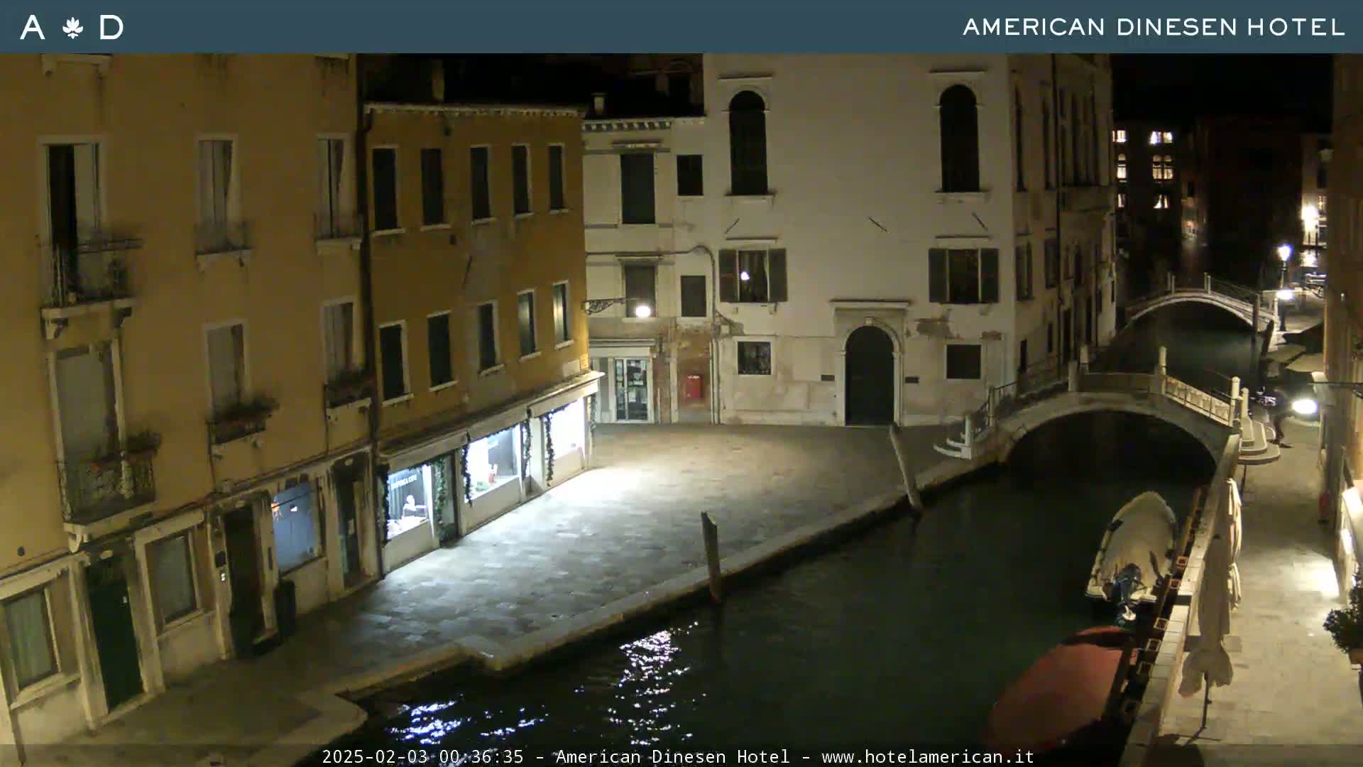 A nighttime view of a canal in Venice, showing several buildings lining the waterway, a small bridge, and a boat moored along the embankment.