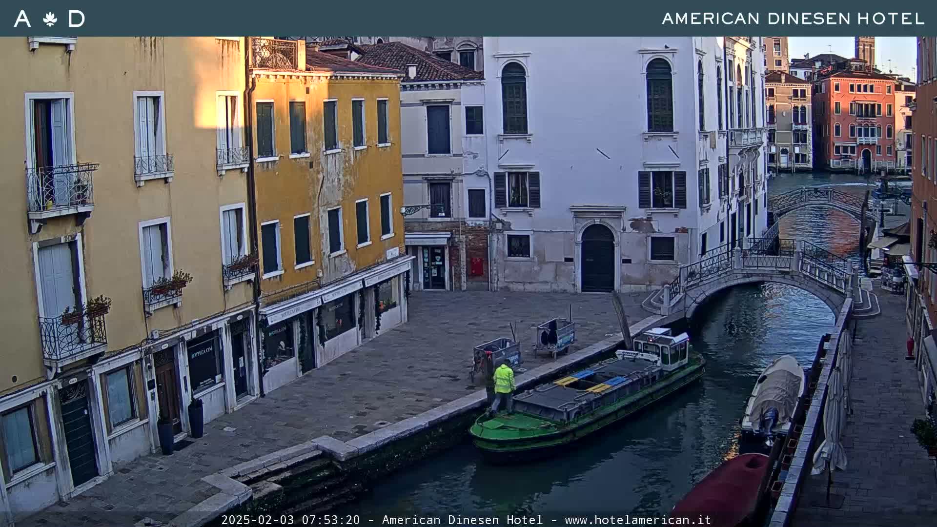 A calm canal in Venice, Italy, is seen at sunrise, with colorful buildings lining its banks and a small green boat docked along the quay.