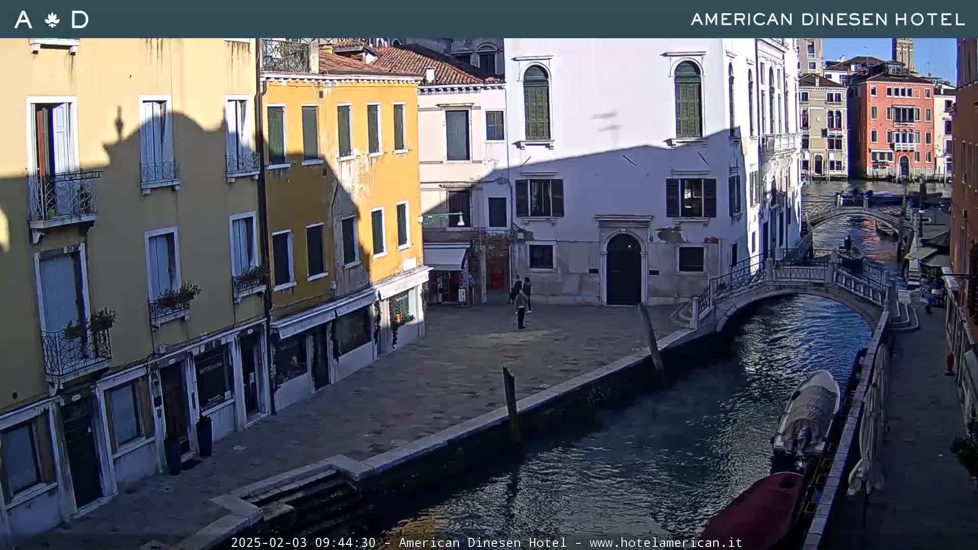 A sunny view of a canal in Venice, Italy, shows colorful buildings lining the waterway, with a bridge and several small boats.