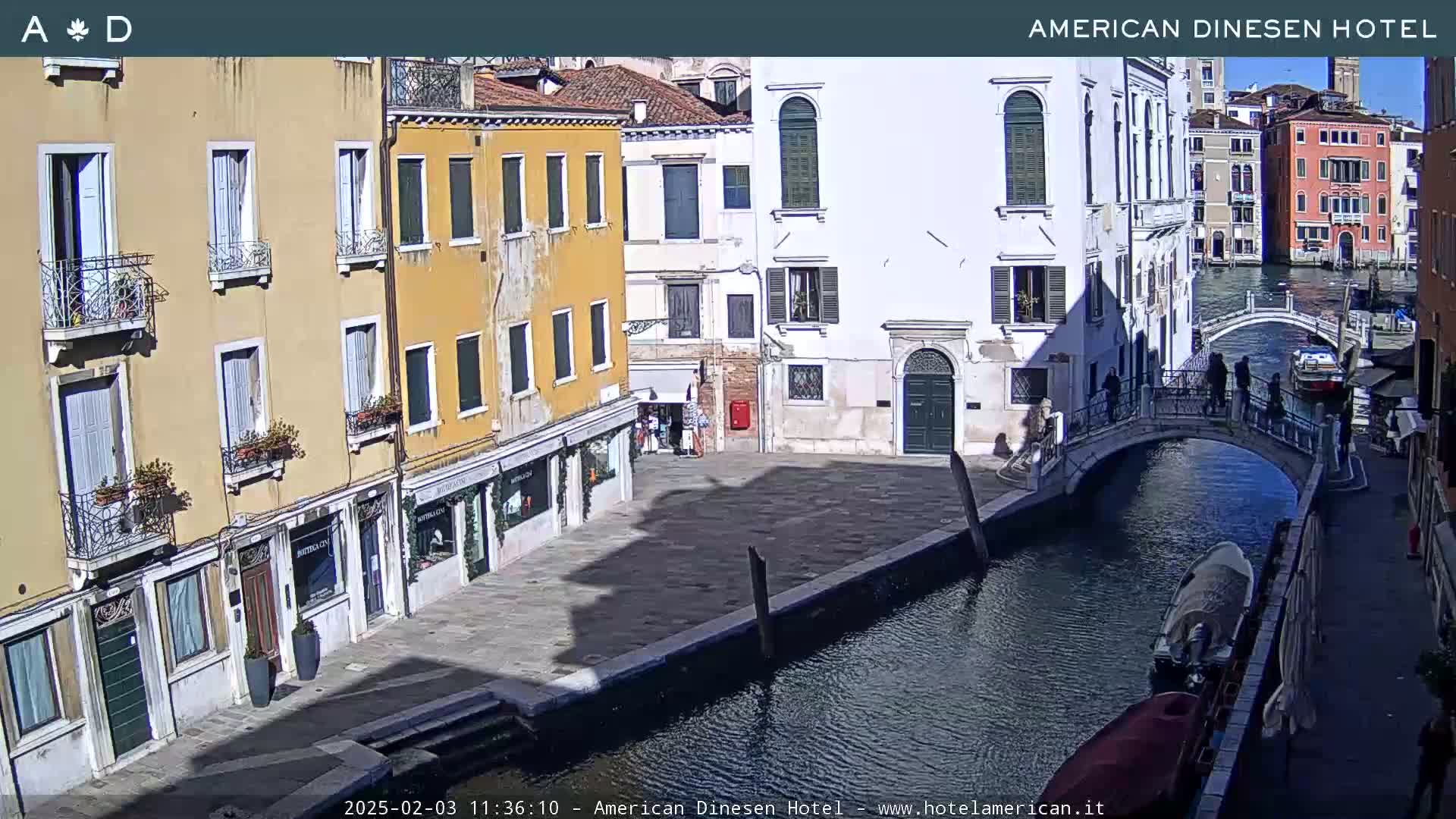 A sunny view of a canal in Venice, Italy, with several buildings lining the waterway and a small bridge with pedestrians.