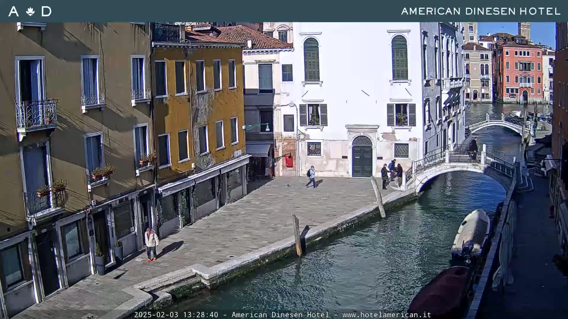A sunny view of a canal in Venice, Italy, with several buildings lining the waterway and a small bridge spanning the canal.