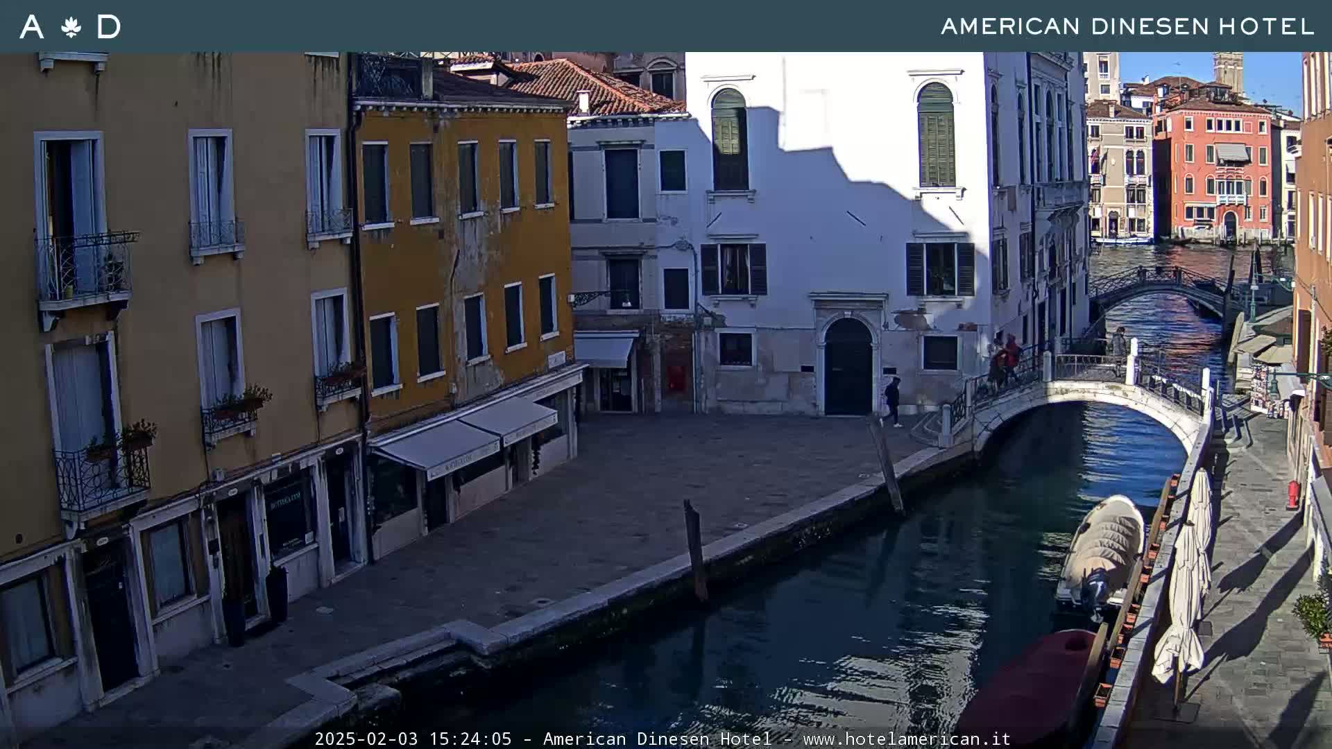 A sunny Venetian canal scene shows several multi-story buildings, a pedestrian bridge, and a small boat on the water.