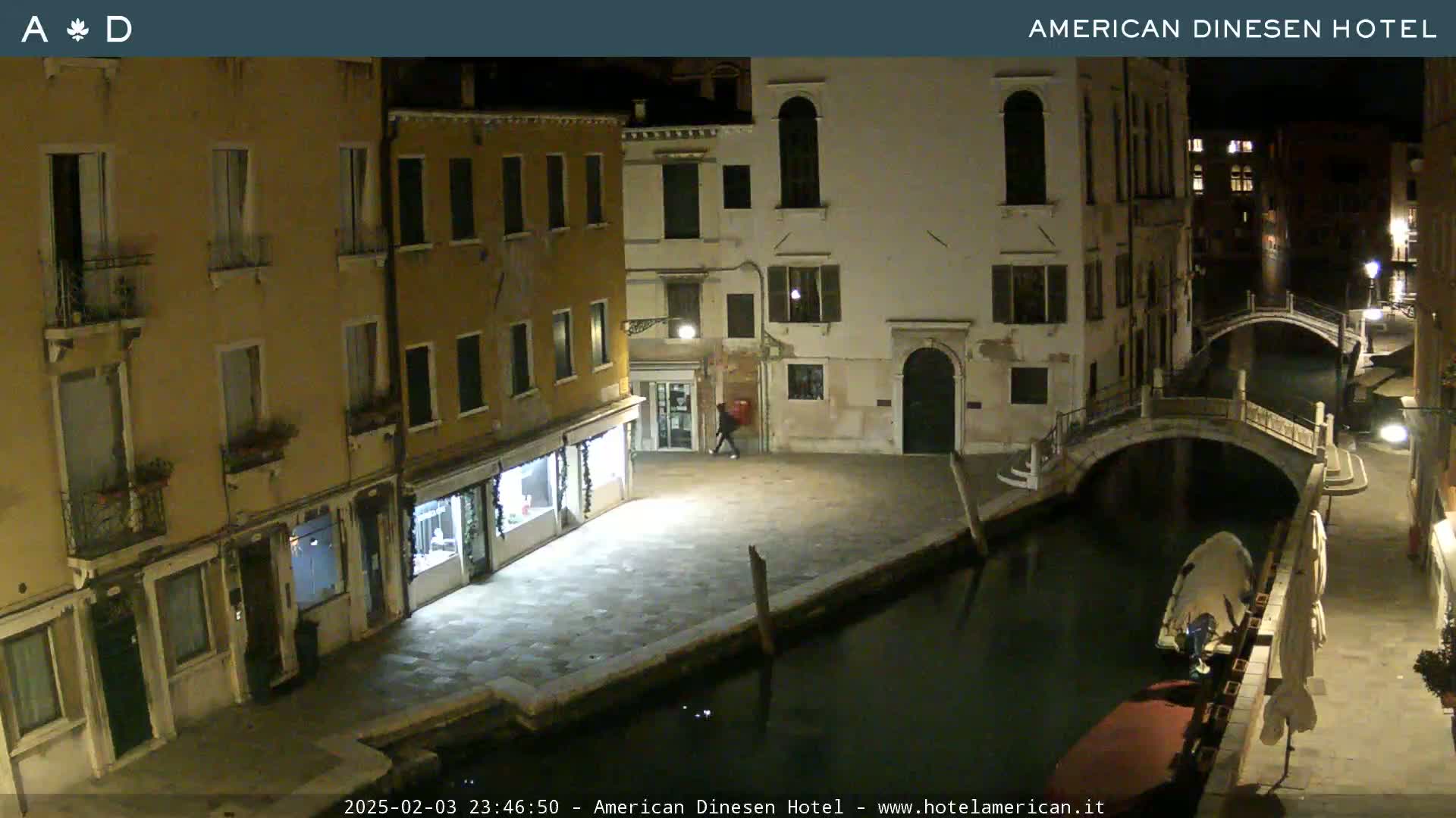 A nighttime view of a canal in Venice, Italy, showing several buildings along the waterway and a small bridge, with a person walking in a nearby square.