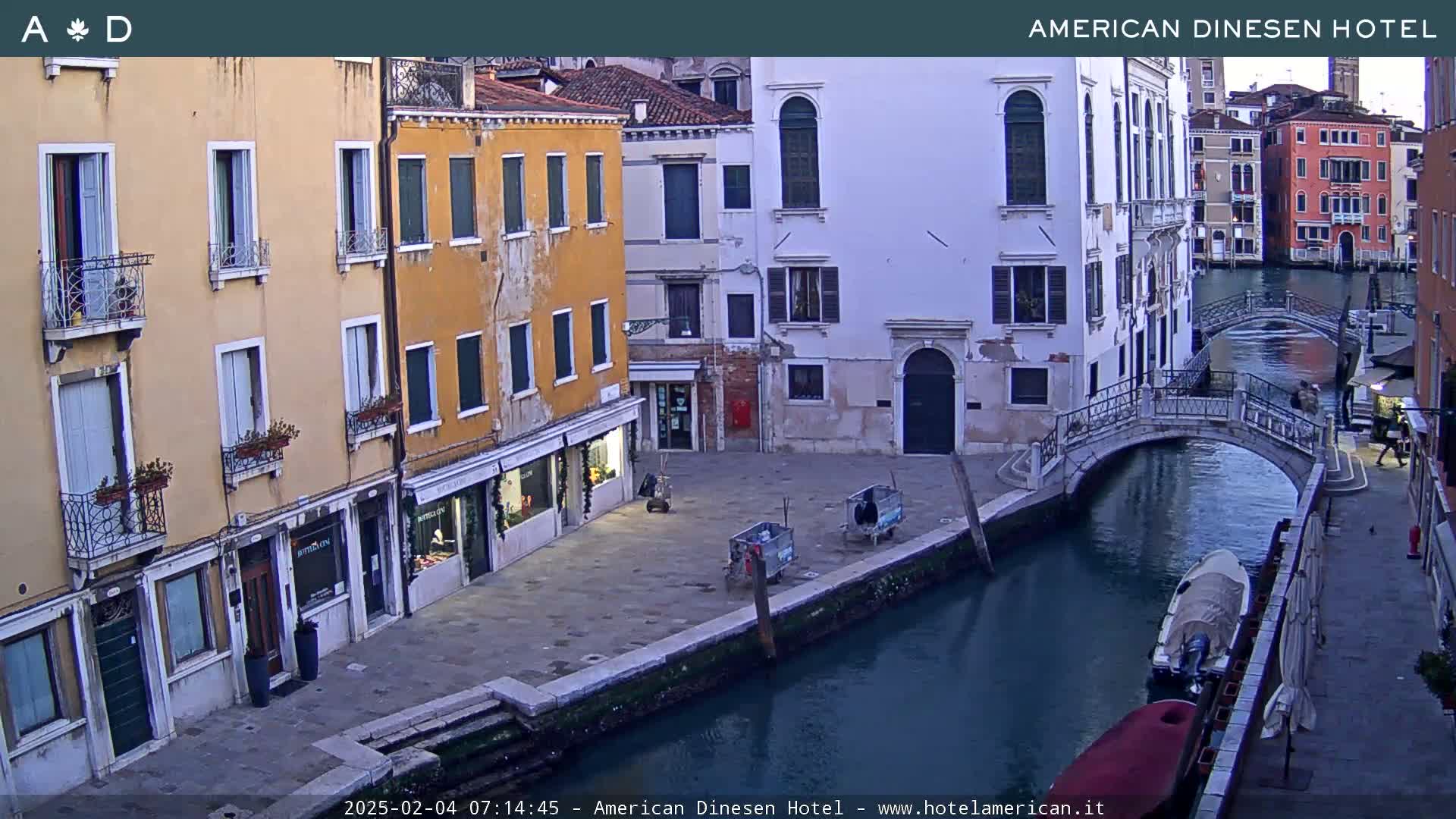 A canal in Venice, Italy, at dusk, shows several buildings lining the waterway, with a small bridge crossing the canal and a few boats moored alongside.