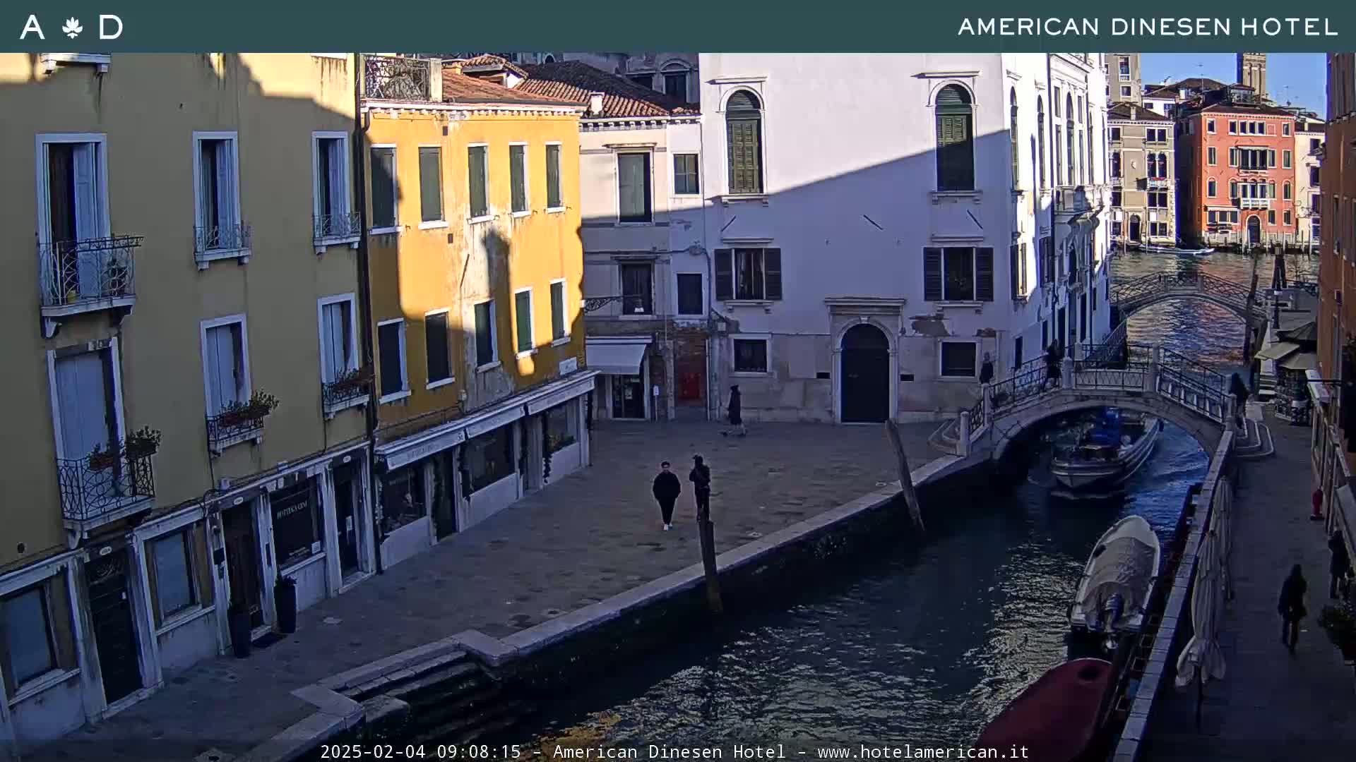 A sunny Venetian canal scene shows colorful buildings lining the waterway, a small bridge, and several boats.