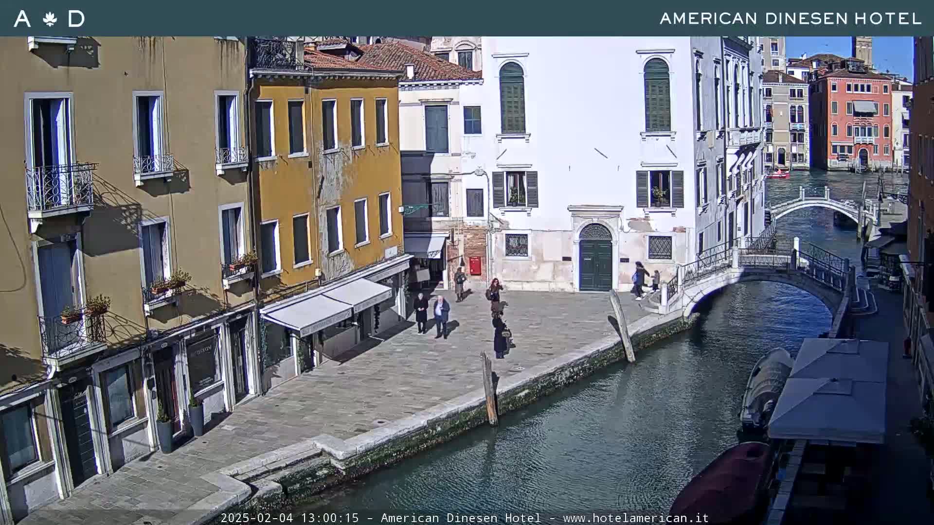 A sunny day in Venice shows several people walking along a canal-side walkway near buildings and a small bridge.