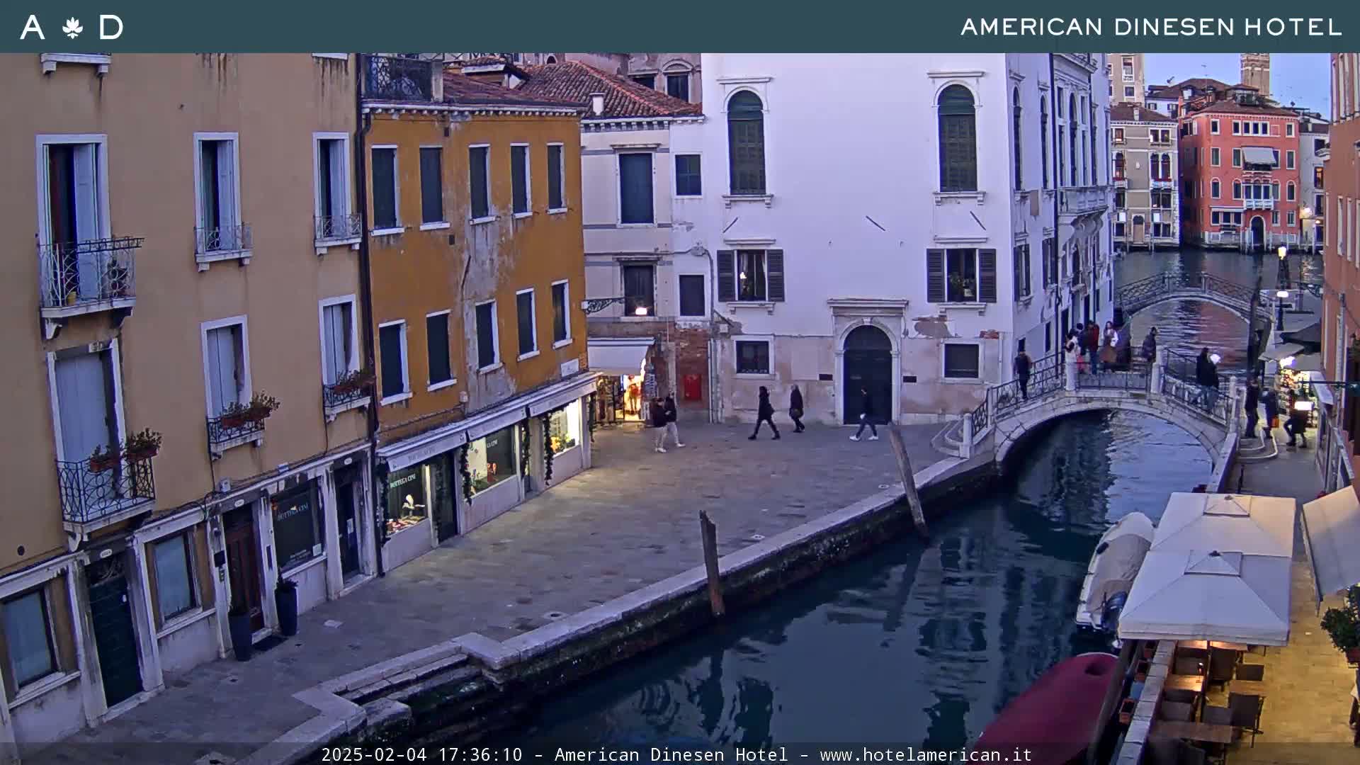 A canal in Venice, Italy at dusk, shows several pedestrians walking along a waterfront path and a small bridge crossing over a calm canal, with buildings lining the waterway.