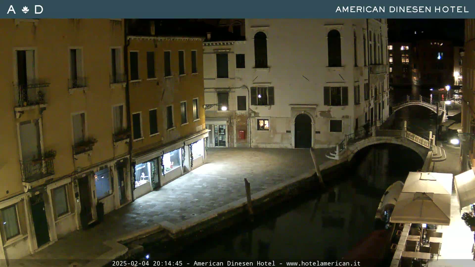 A nighttime view of a canal in Venice, showing several buildings along its edge, a bridge arching over the water, and a small outdoor seating area.