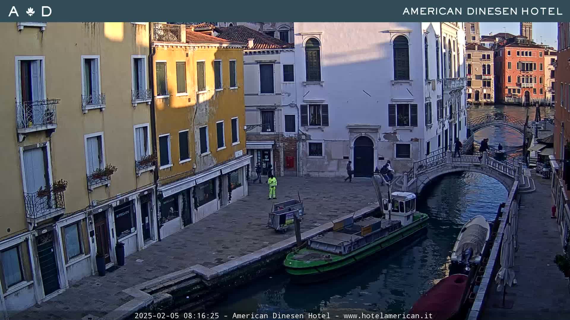A Venetian canal scene at dusk shows several buildings lining the waterway, a small bridge, and a few pedestrians and a small work boat.