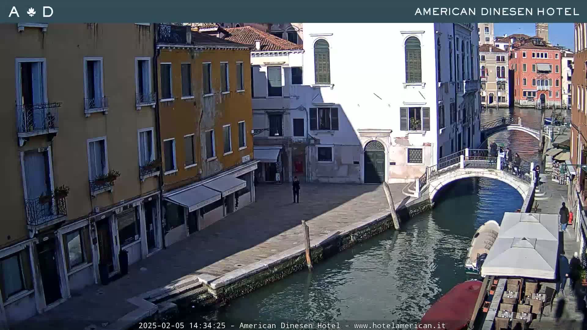 A sunny view of a canal in Venice, Italy, shows several buildings, a pedestrian bridge, and a few people walking along the waterway.