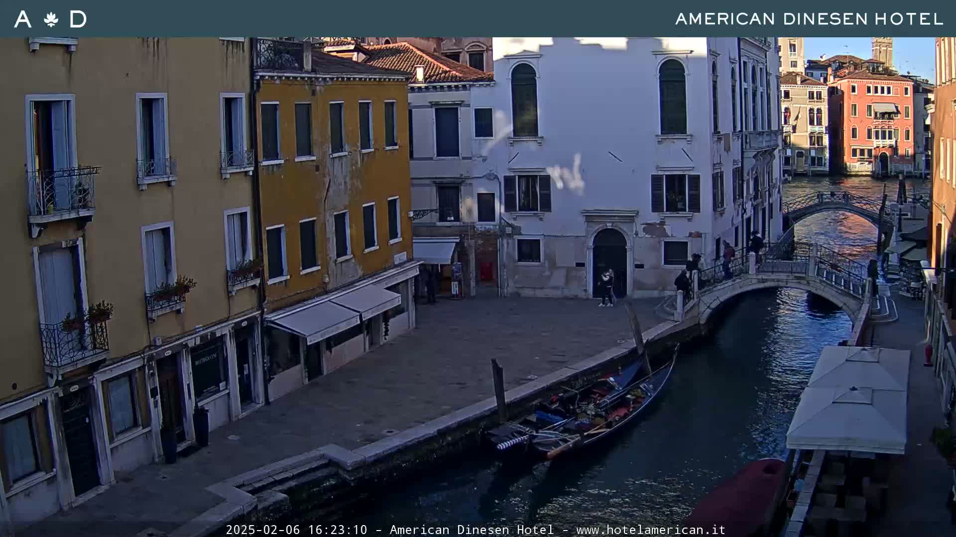 A gondola is moored beside a canal in Venice, under a partly cloudy sky, with several buildings lining the waterway and a small bridge visible.