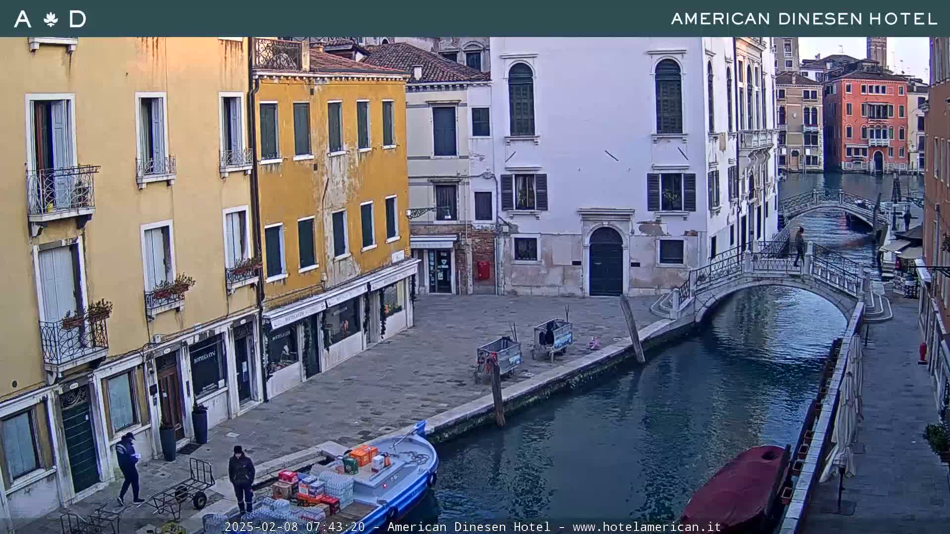 A canal in Venice, under a partly cloudy sky, shows several buildings along its banks, a small boat docked, and a pedestrian bridge.