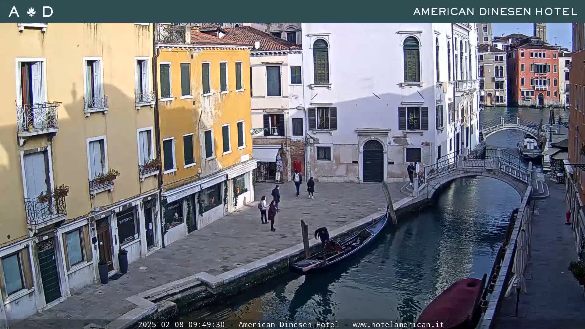 A gondola is moored by a canal in Venice, Italy, on a sunny day, with several people standing on a stone walkway next to brightly colored buildings.
