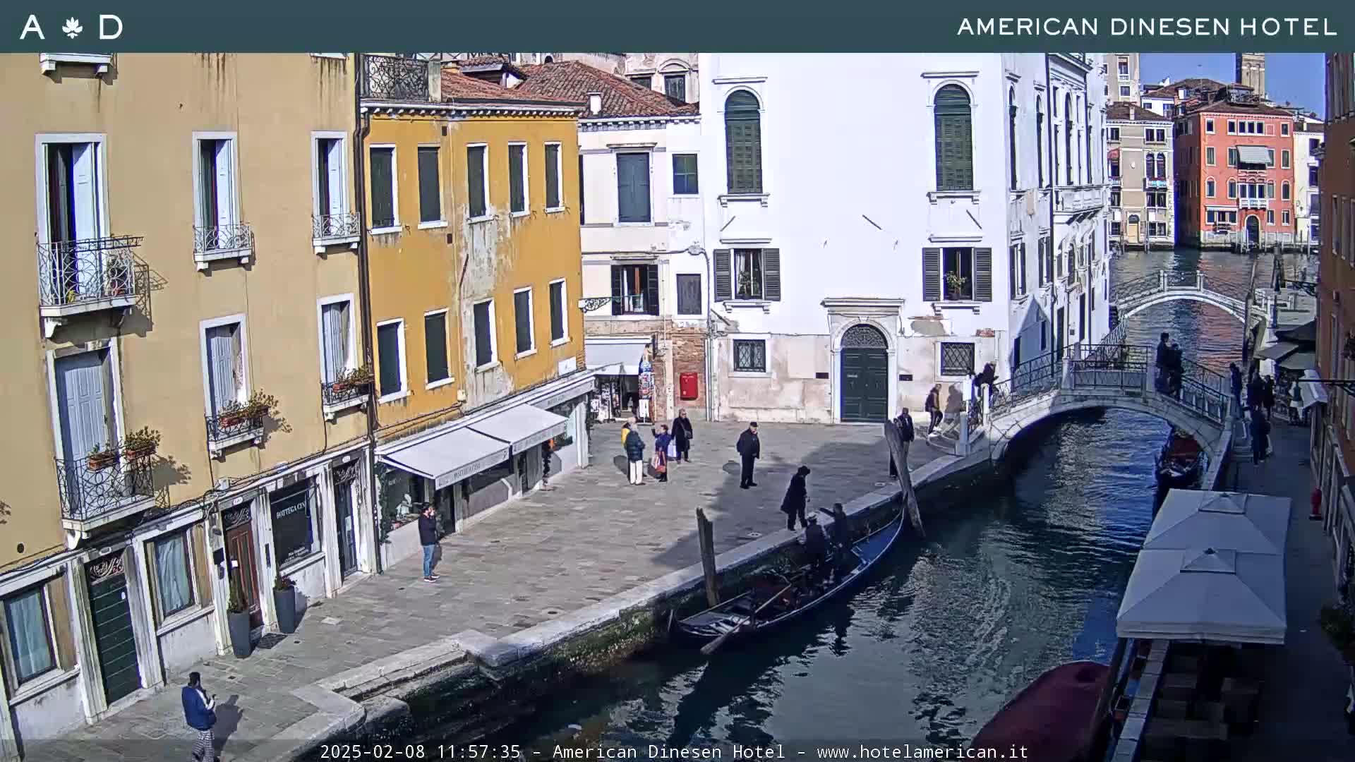A sunny view of a Venetian canal with gondolas, pedestrians on a bridge and cobblestone streets, and several buildings lining the waterway.