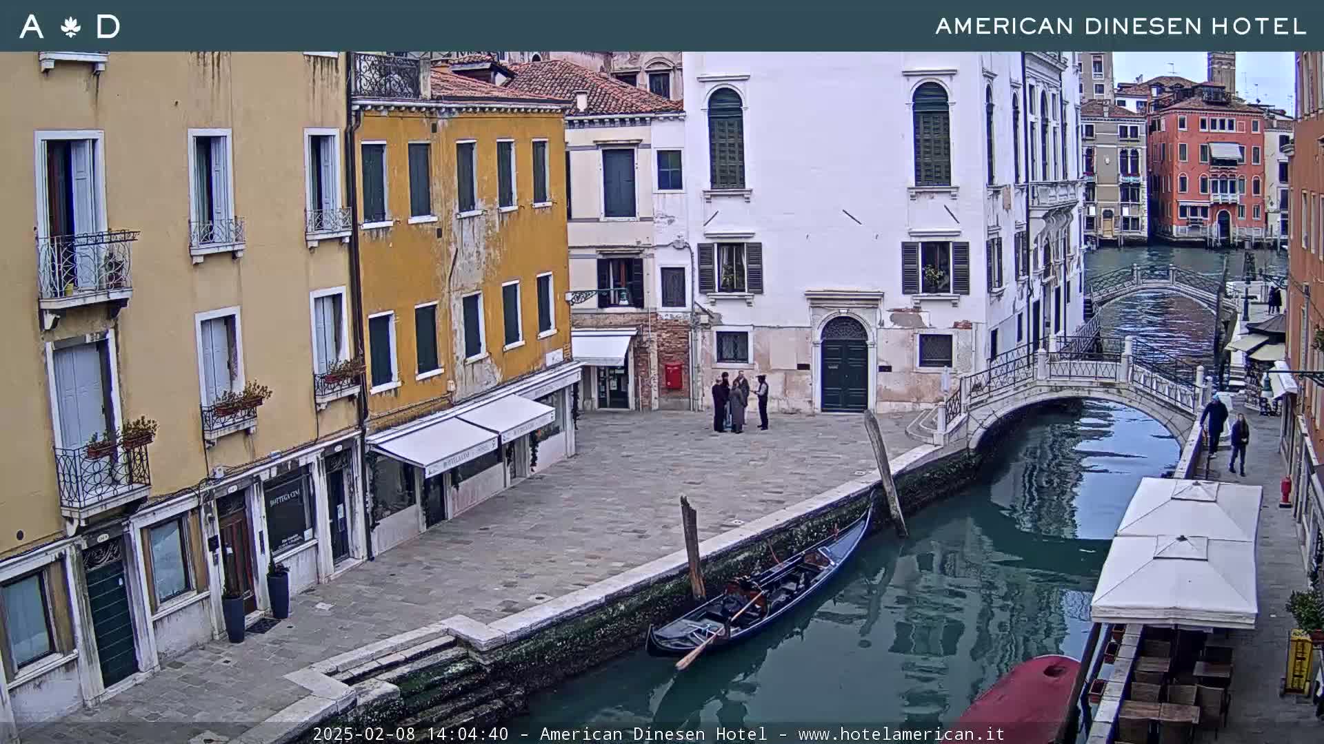 A gondola is moored alongside a canal in Venice, Italy, under a cloudy sky, with several buildings and a small footbridge visible.