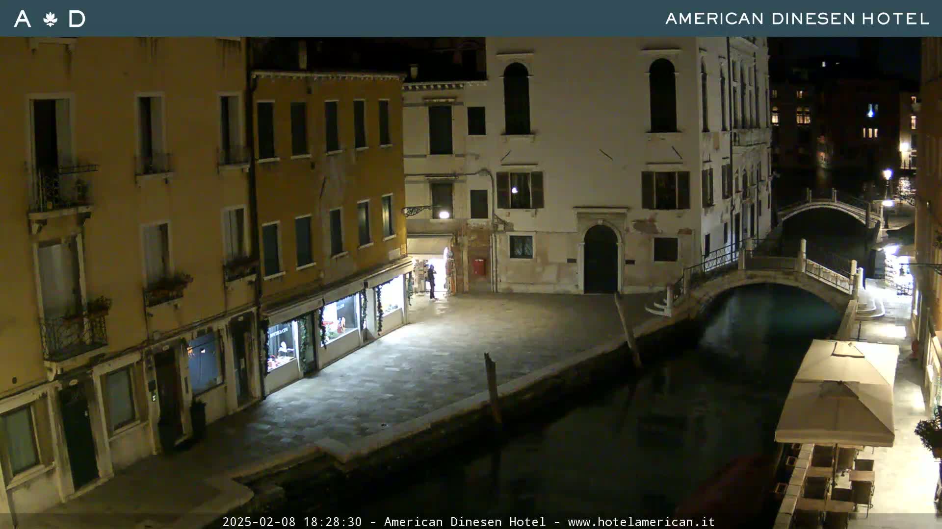 A nighttime view of a canal in Venice, Italy, shows several buildings lining the waterway, a small bridge crossing the canal, and a few people on the street.