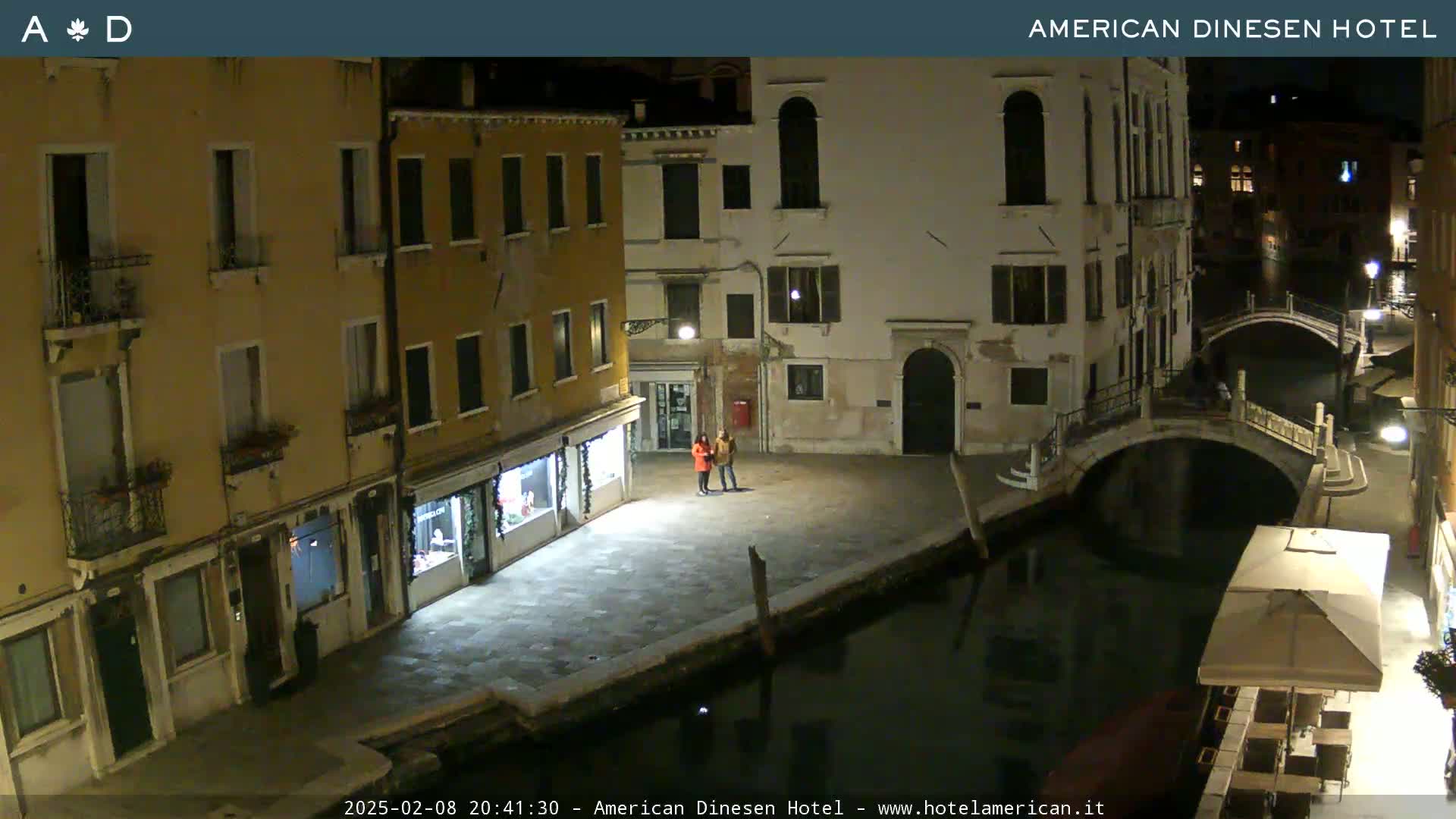 A nighttime view of a canal in Venice, Italy, shows several buildings lining the waterway, a small bridge crossing the canal, and two people standing in a plaza.