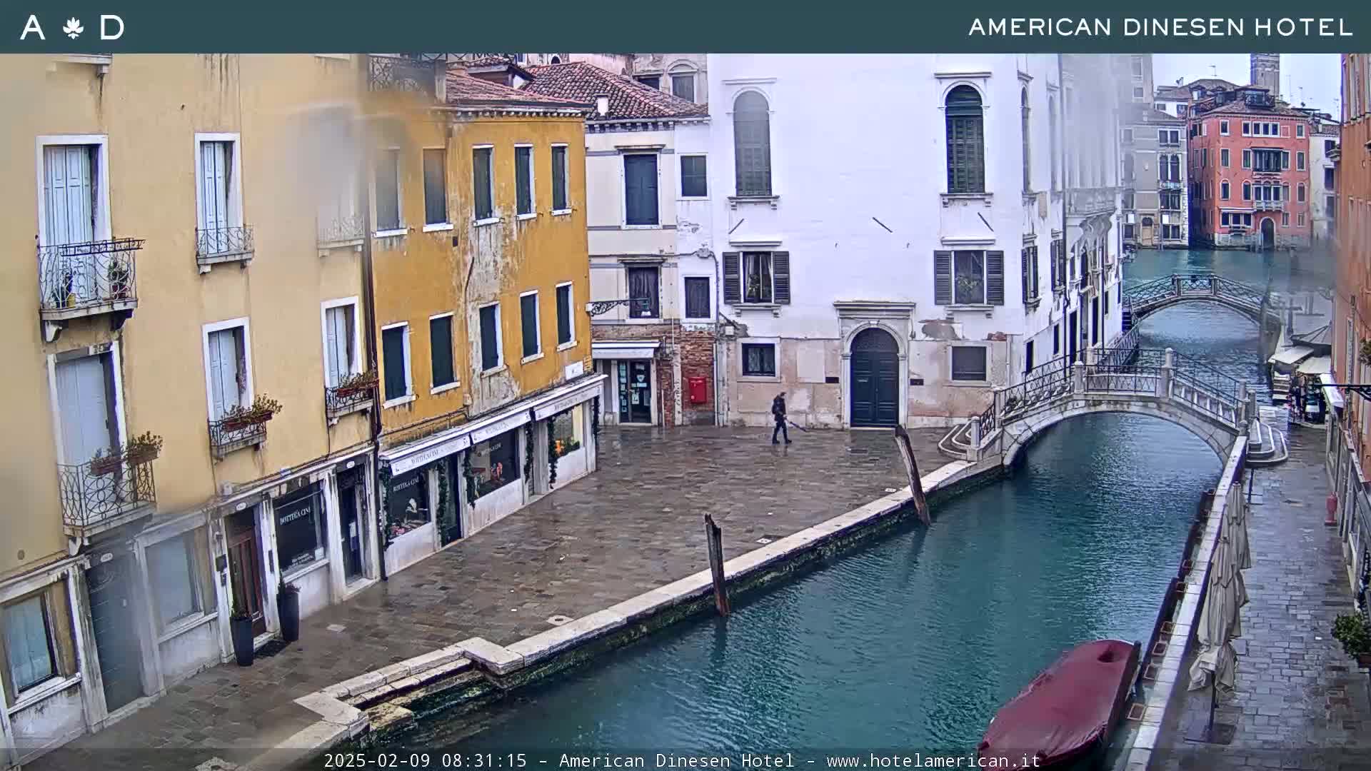 A canal in Venice, Italy, with overcast skies, shows two bridges and several buildings lining its banks, one of which has a person sweeping near its entrance.