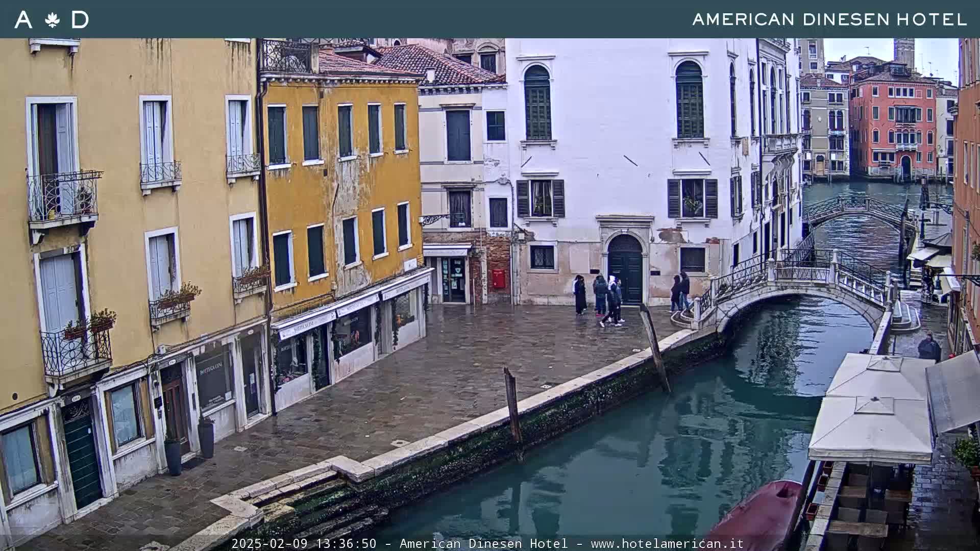 A view of a canal in Venice, Italy, on an overcast day, shows several buildings lining the waterway, a small bridge crossing the canal, and a few people standing on a stone walkway.