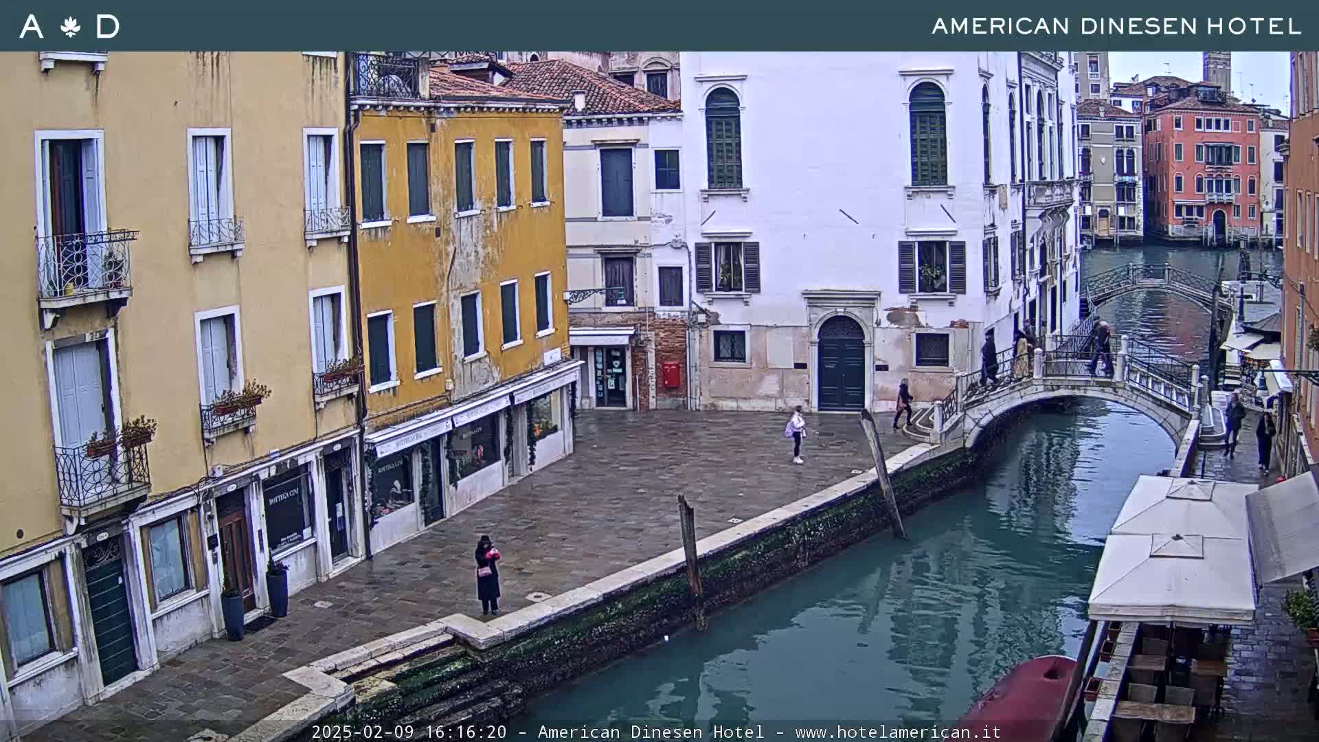 A view of a canal in Venice, Italy, on an overcast day, shows several buildings lining the waterway, with a small footbridge crossing the canal and a few pedestrians visible.