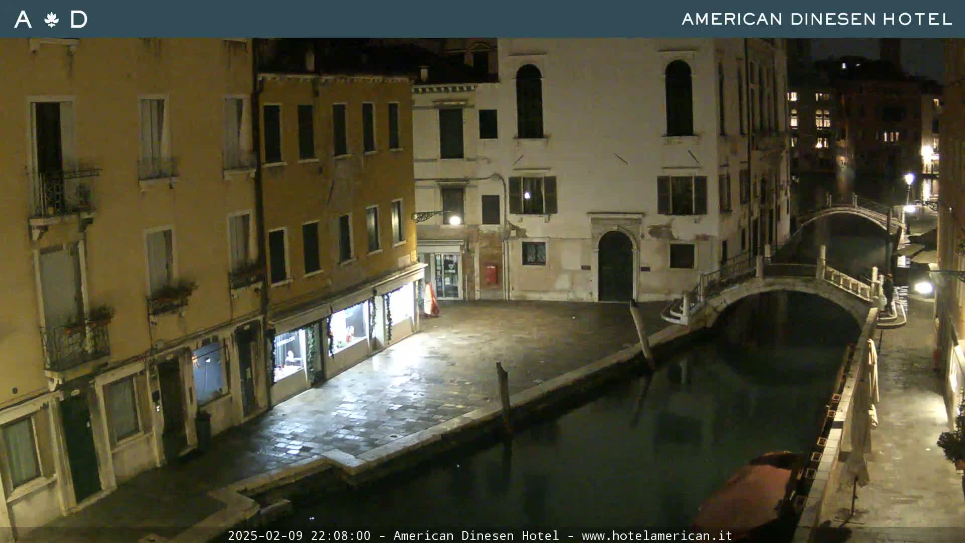 A nighttime view of a canal in Venice, Italy, shows several buildings lining the waterway, a stone bridge spanning the canal, and a small boat moored along the bank.