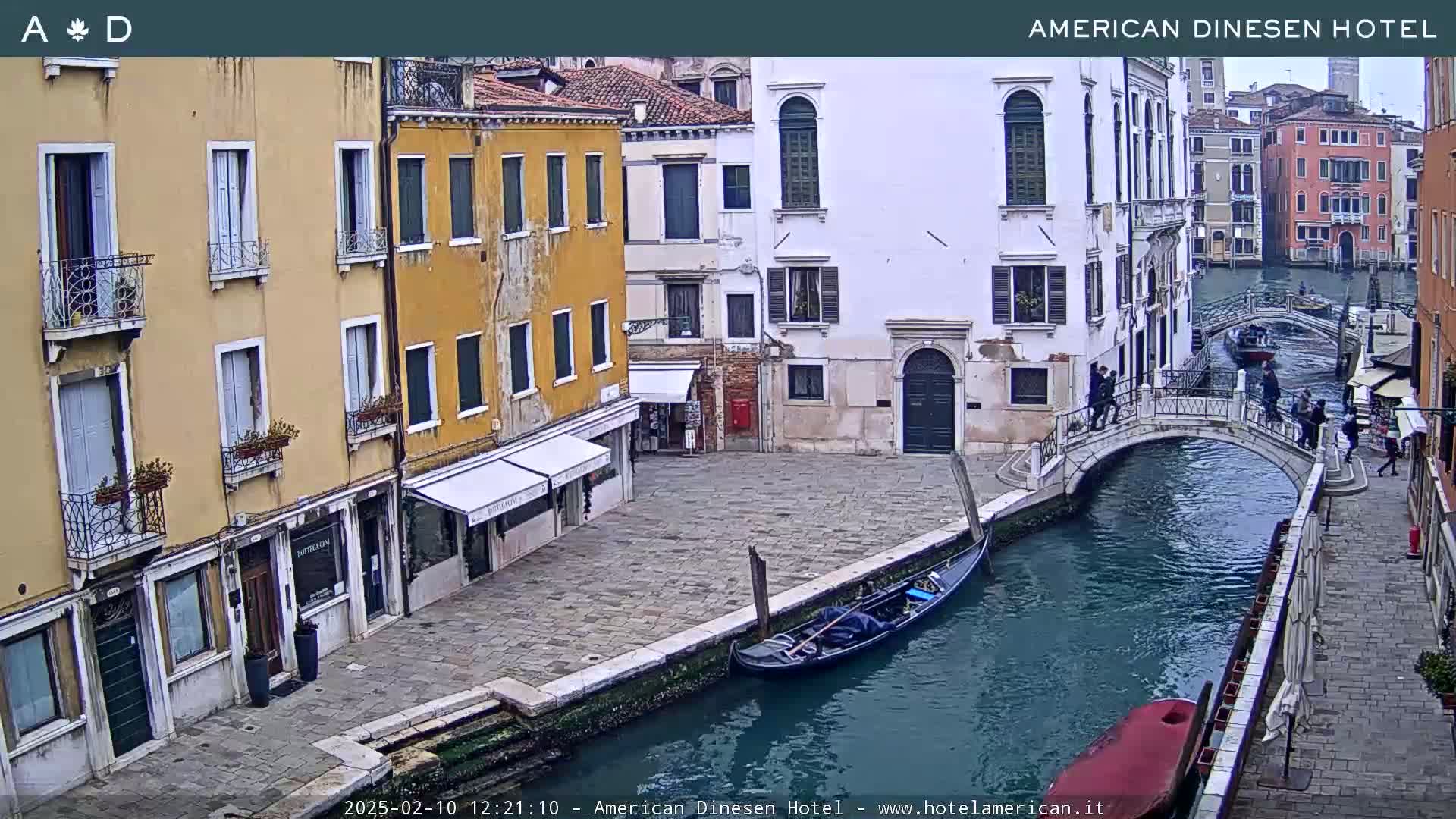 A gondola floats on a canal in Venice, Italy, next to a stone bridge where pedestrians are walking, with several multi-story buildings lining the waterway.