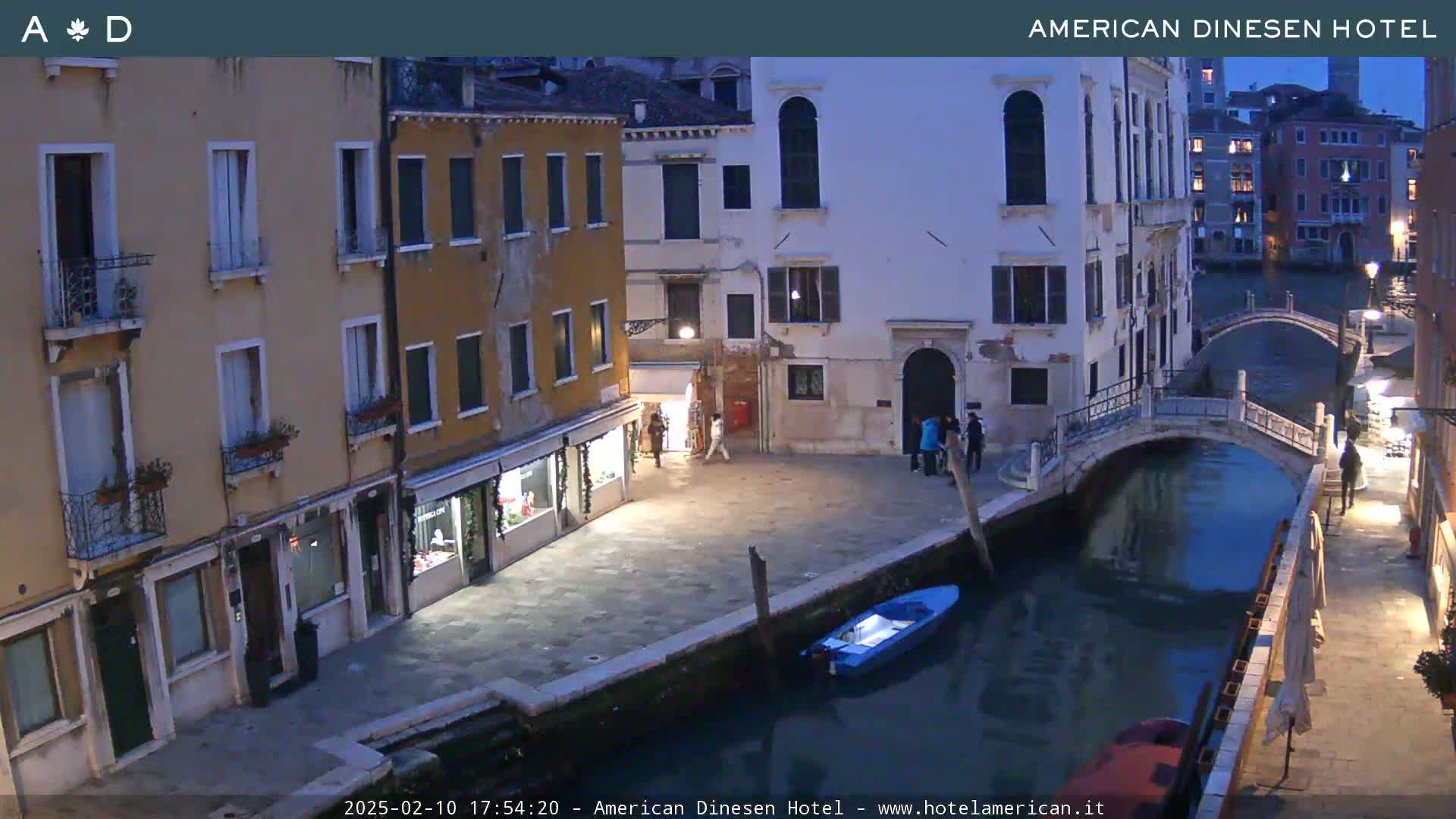A nighttime view of a canal in Venice, showing several buildings and a small bridge, with a few people walking along the canal's edge.