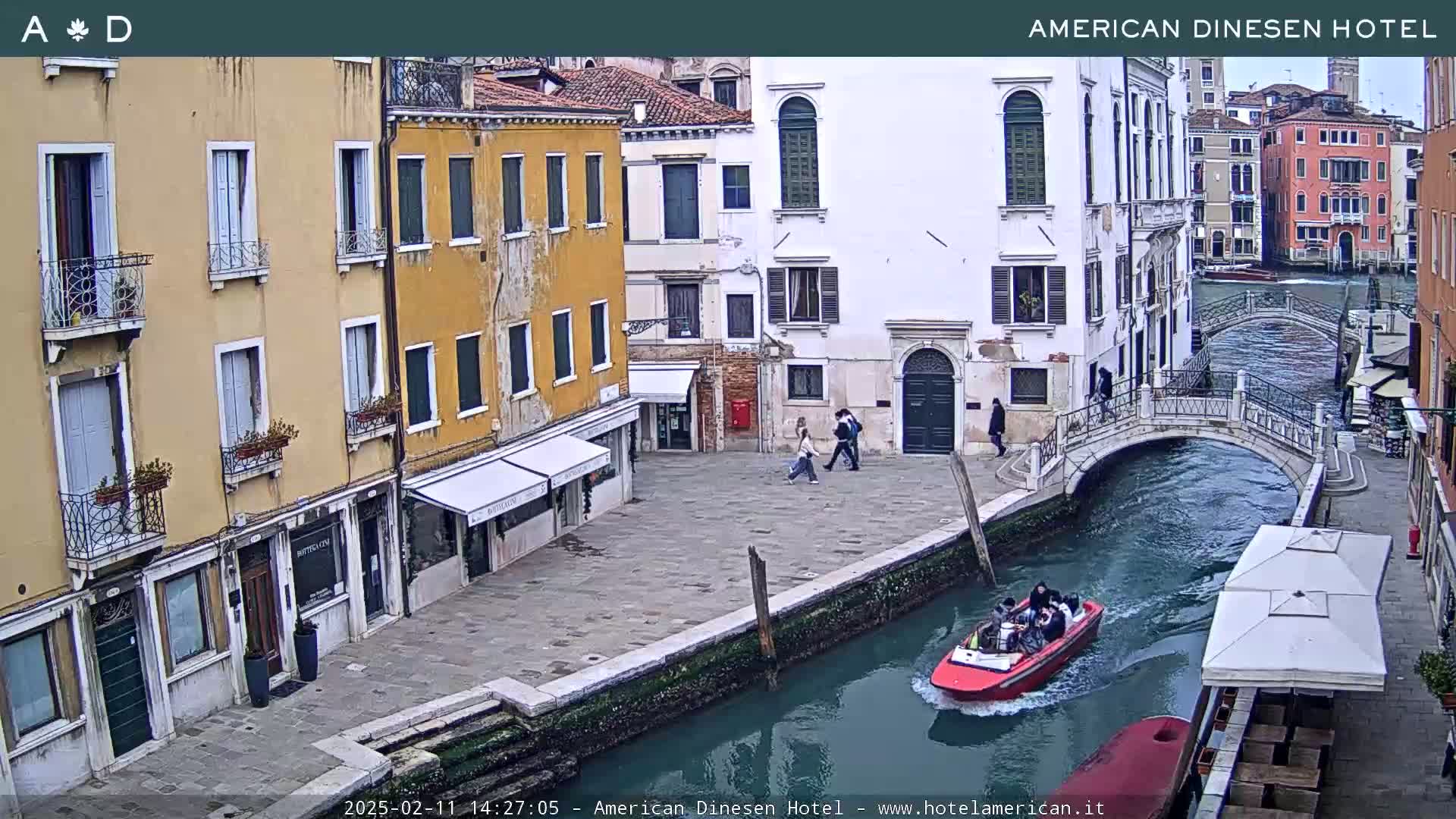 A waterway in Venice, Italy, shows a red motorboat carrying passengers passing under a small bridge near several buildings on a cloudy day.
