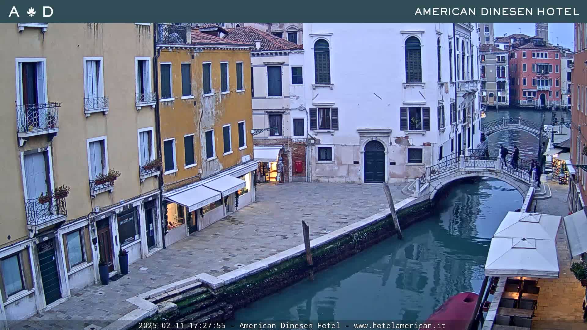 A narrow canal in Venice, under a cloudy sky, is spanned by a footbridge with several pedestrians crossing it, and lined with old buildings.