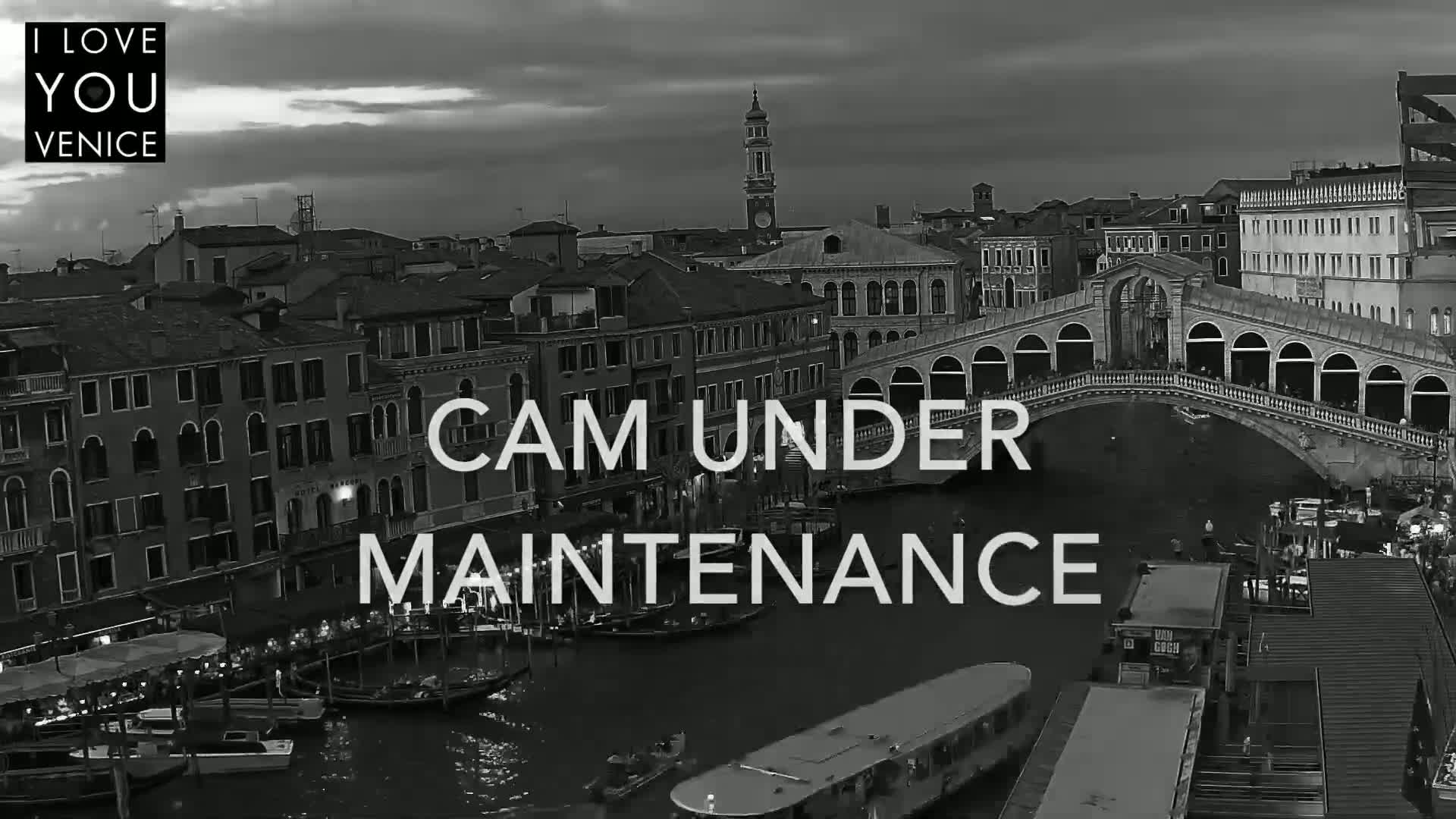 A black and white, nighttime view of a canal in Venice, Italy, shows boats moored along the waterway, a bridge arching over the canal, and buildings lining the banks under a cloudy sky.