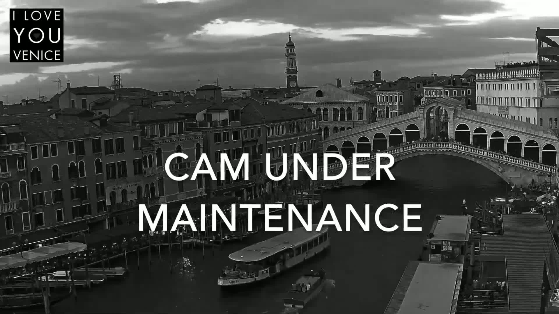 A black and white, high-angle view shows a canal in Venice, Italy, under a partly cloudy sky, with a bridge and many buildings visible.