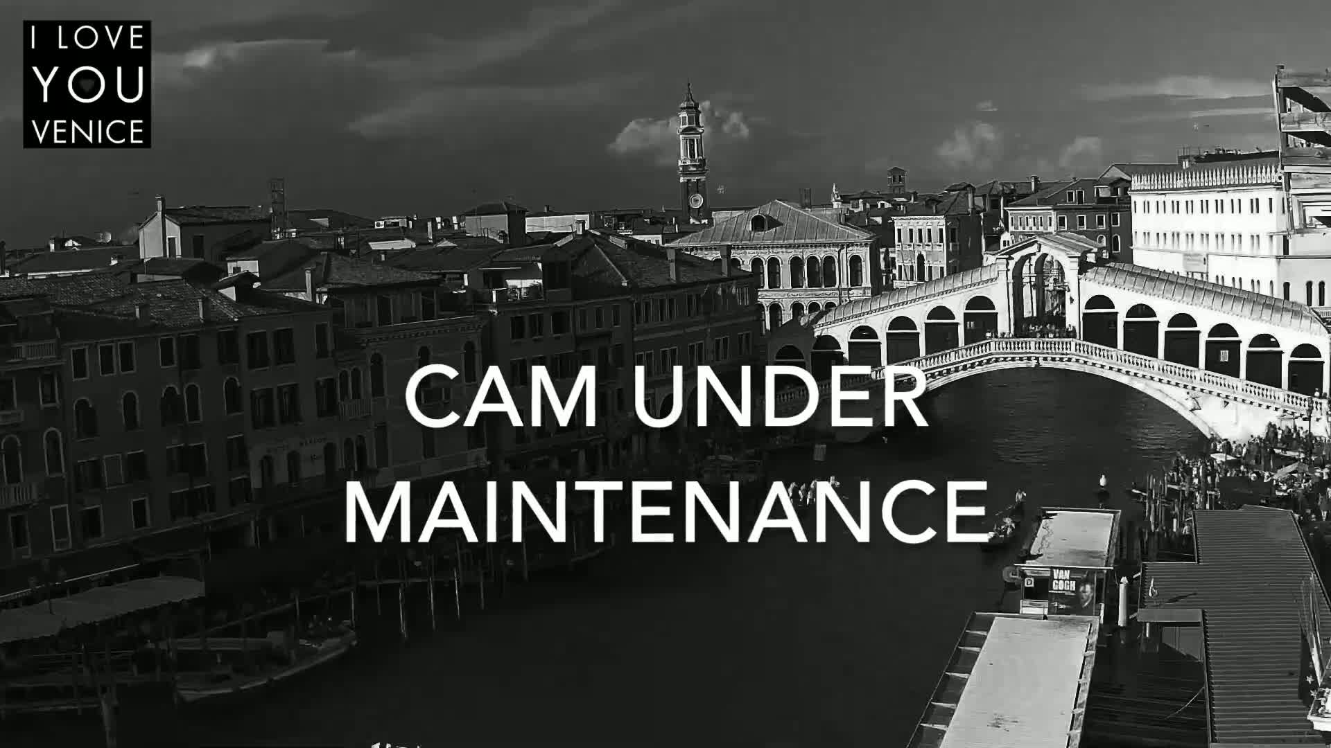 A black and white, high-angle view shows a bridge over a canal in Venice, with many buildings lining the waterway under a partly cloudy sky.