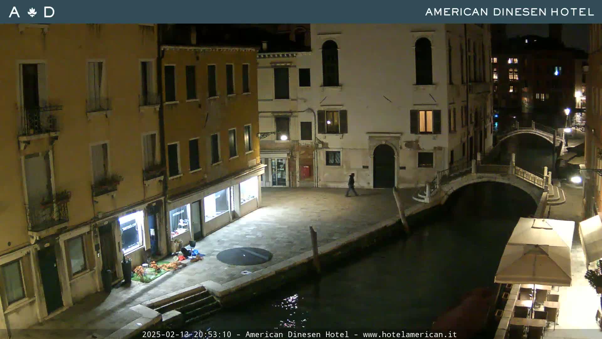 A nighttime view of a canal in Venice, with buildings lining the waterway and a few pedestrians visible.