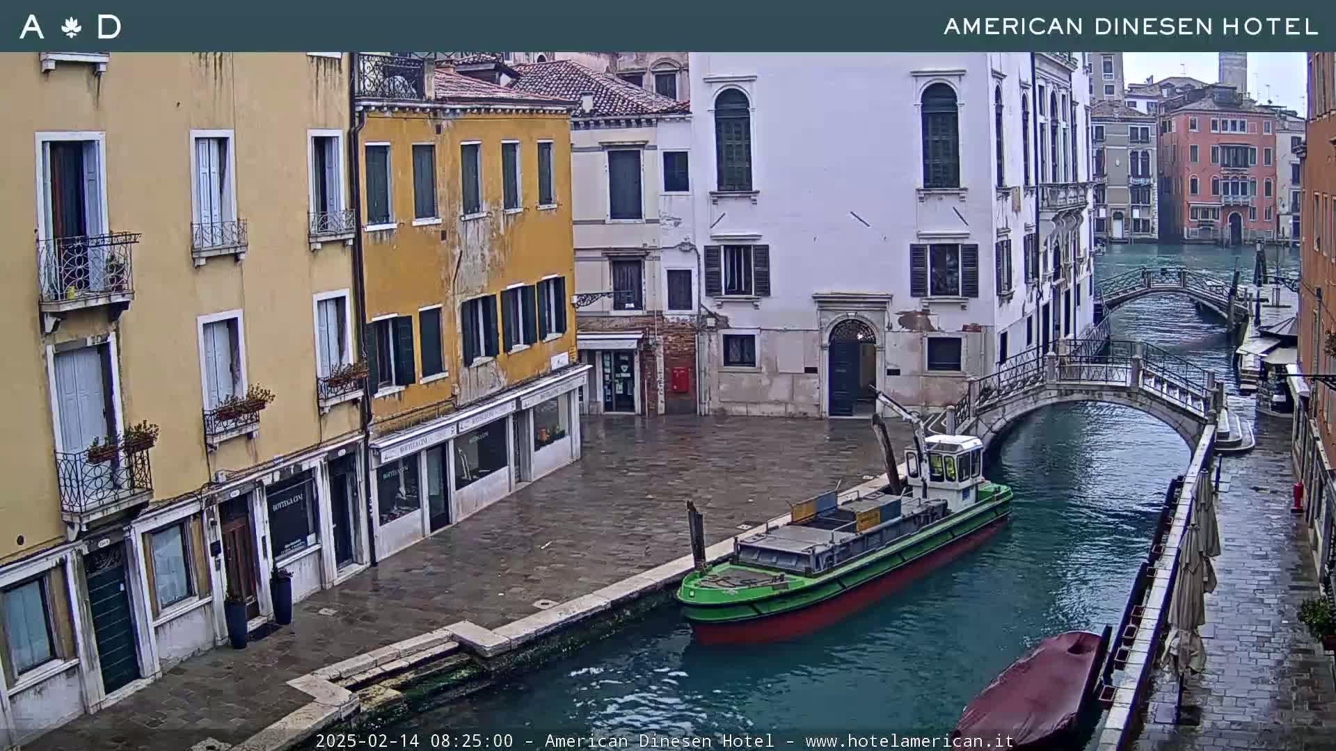 A view of a canal in Venice, Italy, showing several buildings, two small bridges, and a green barge on the water, under overcast skies.